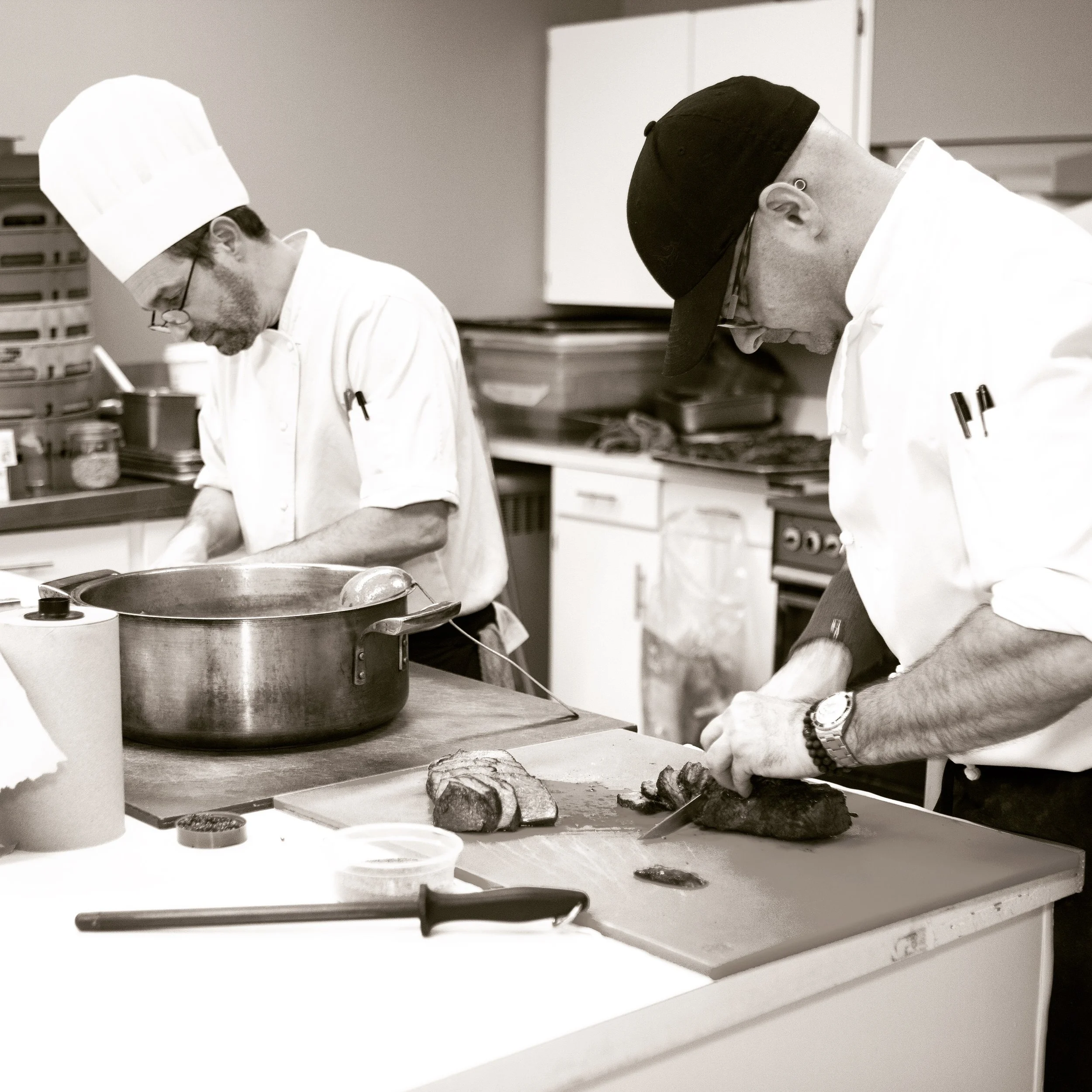 Two chefs in white uniforms and black caps working in a commercial kitchen, preparing food with various cooking tools and ingredients on the table.