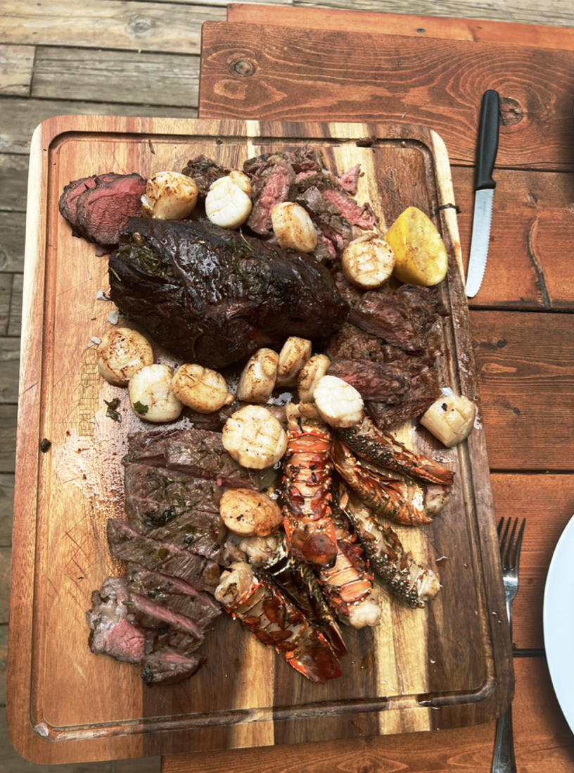 Cut and cooked steak, lobster tails, and roasted scallops on a wooden cutting board.
