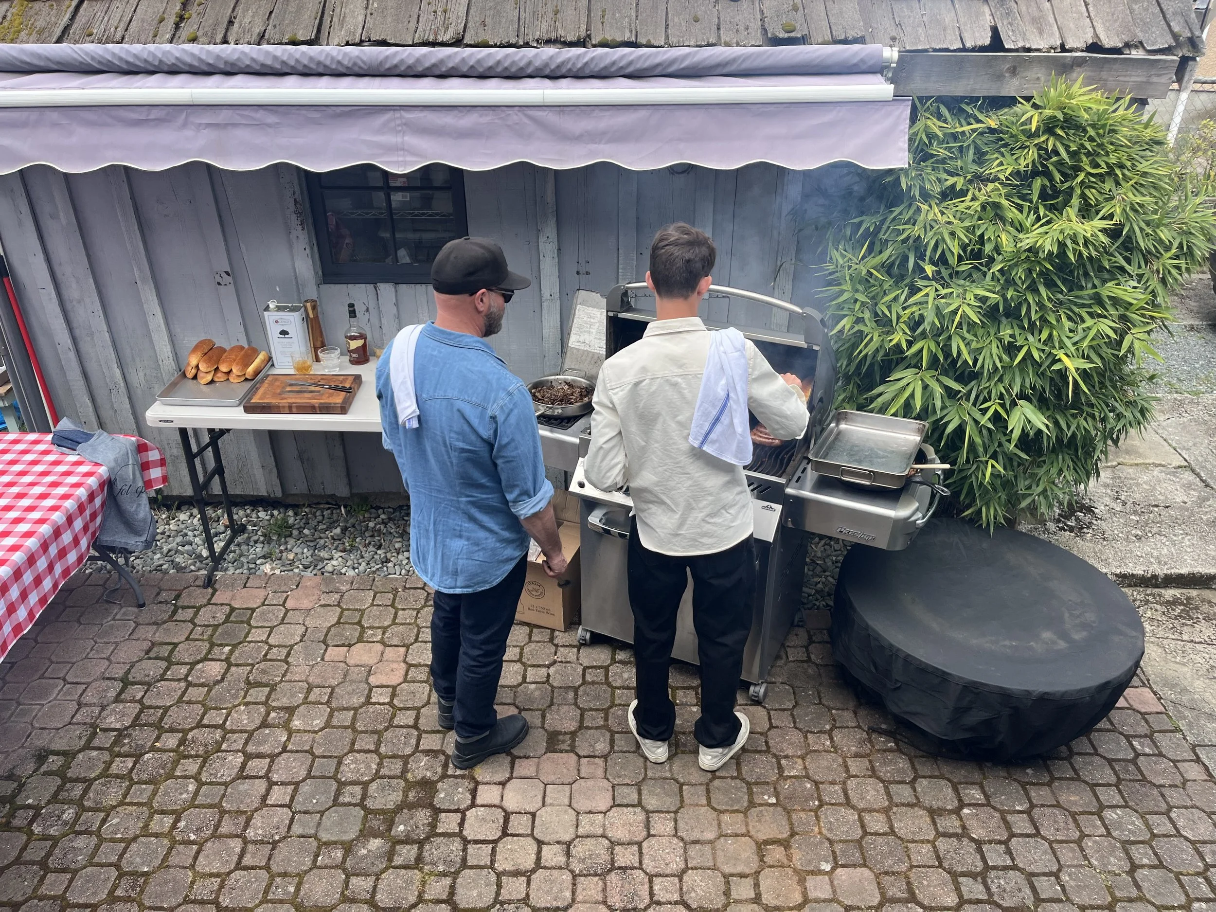 Two men grilling food on a barbecue grill in a backyard, with a wooden wall and green shrubbery in the background. One man is wearing a blue shirt and a black baseball cap, while the other is in a beige jacket with a towel over his shoulder.
