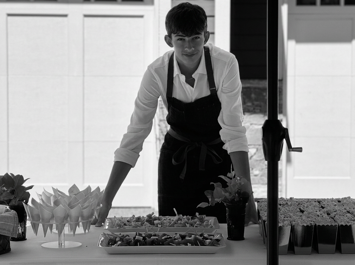 A young man wearing a white shirt and a dark apron stands behind a table with arranged snacks and flowers, smiling at the camera, indoors with a bright background.
