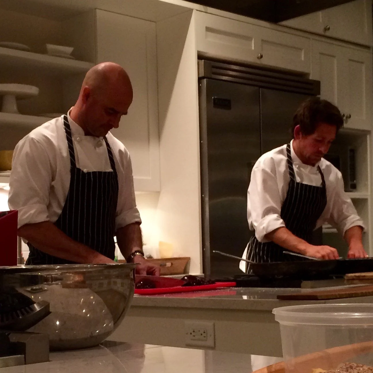 Two male chefs in white shirts and black-and-white striped aprons preparing food in a kitchen.
