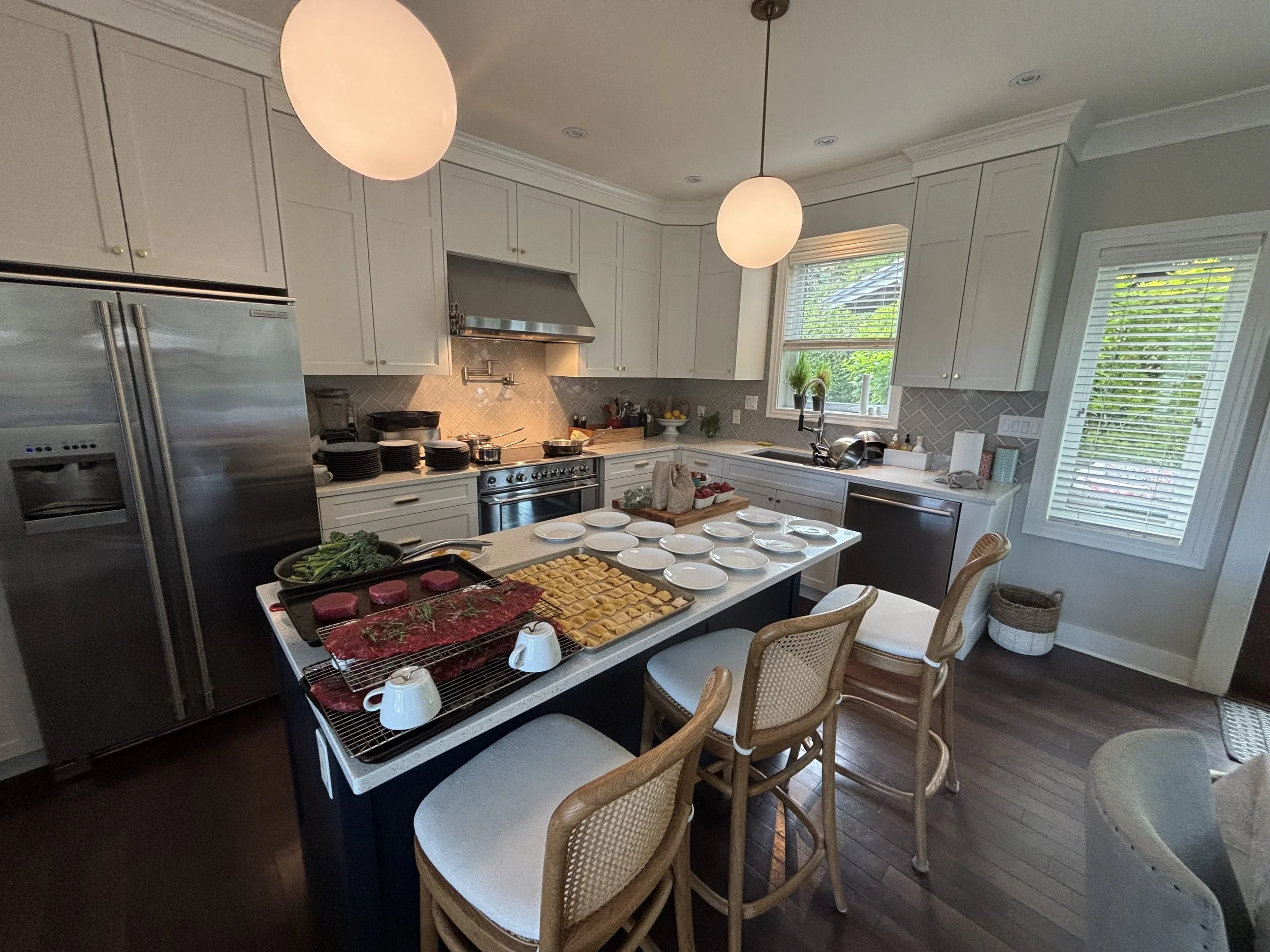 A modern kitchen decorated for a gathering with a large island table filled with plates, cups, and various food items including trays of raw meat and vegetables, surrounded by woven chairs and illuminated by two hanging pendant lights near a window.