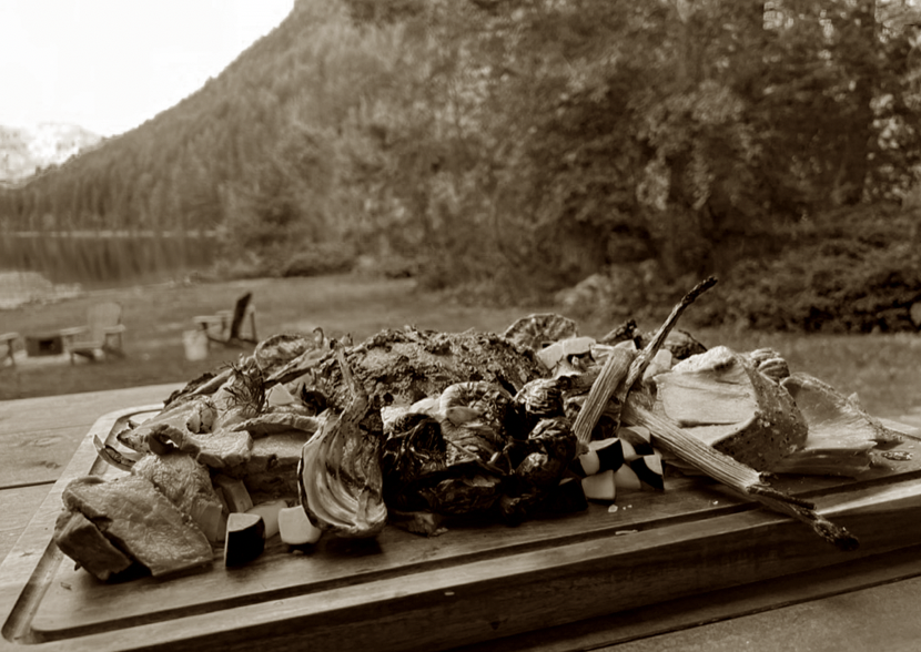 A plate of grilled seafood and vegetables on a wooden board, with a lakeside and forested mountains in the background.