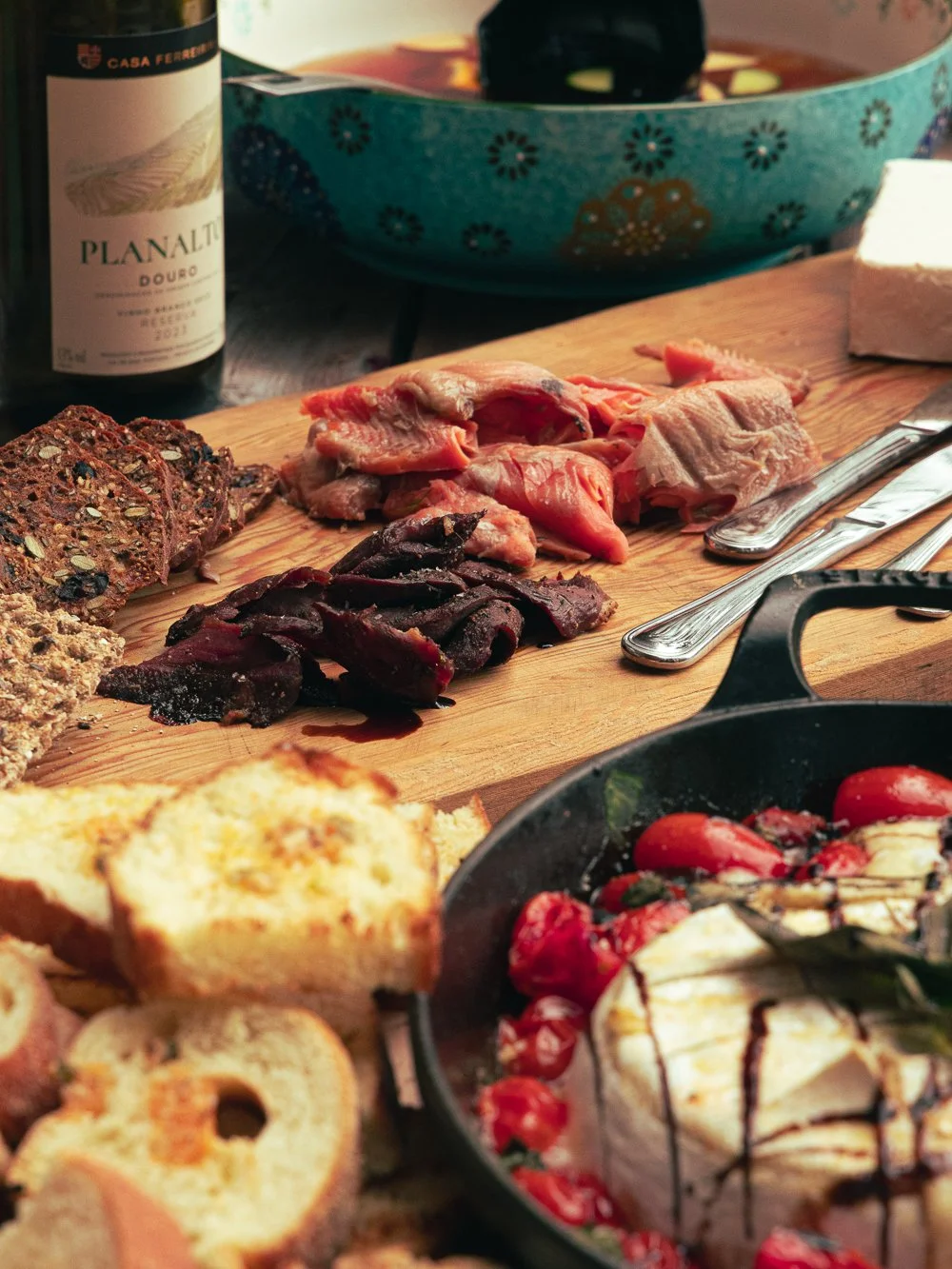 A charcuterie board with sliced meats, a variety of cheeses, and bread with berries and chocolate sauce nearby. A bottle of wine and a bowl with a floral pattern are in the background.