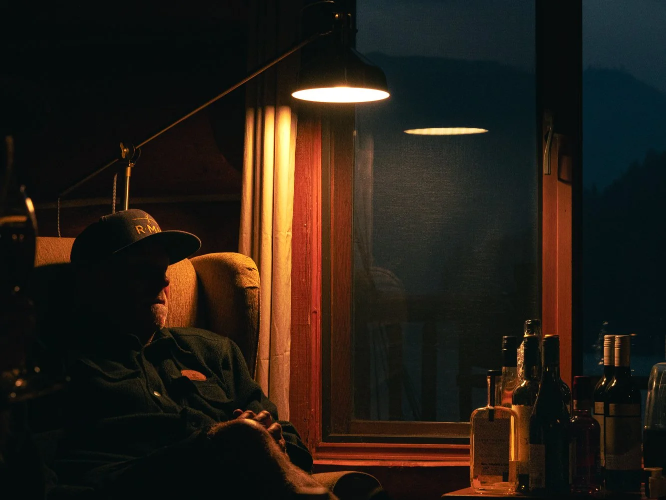 A man sitting in a dimly lit room at night, wearing a cap, with bar bottles on a table nearby, and a window showing a dark landscape outside.