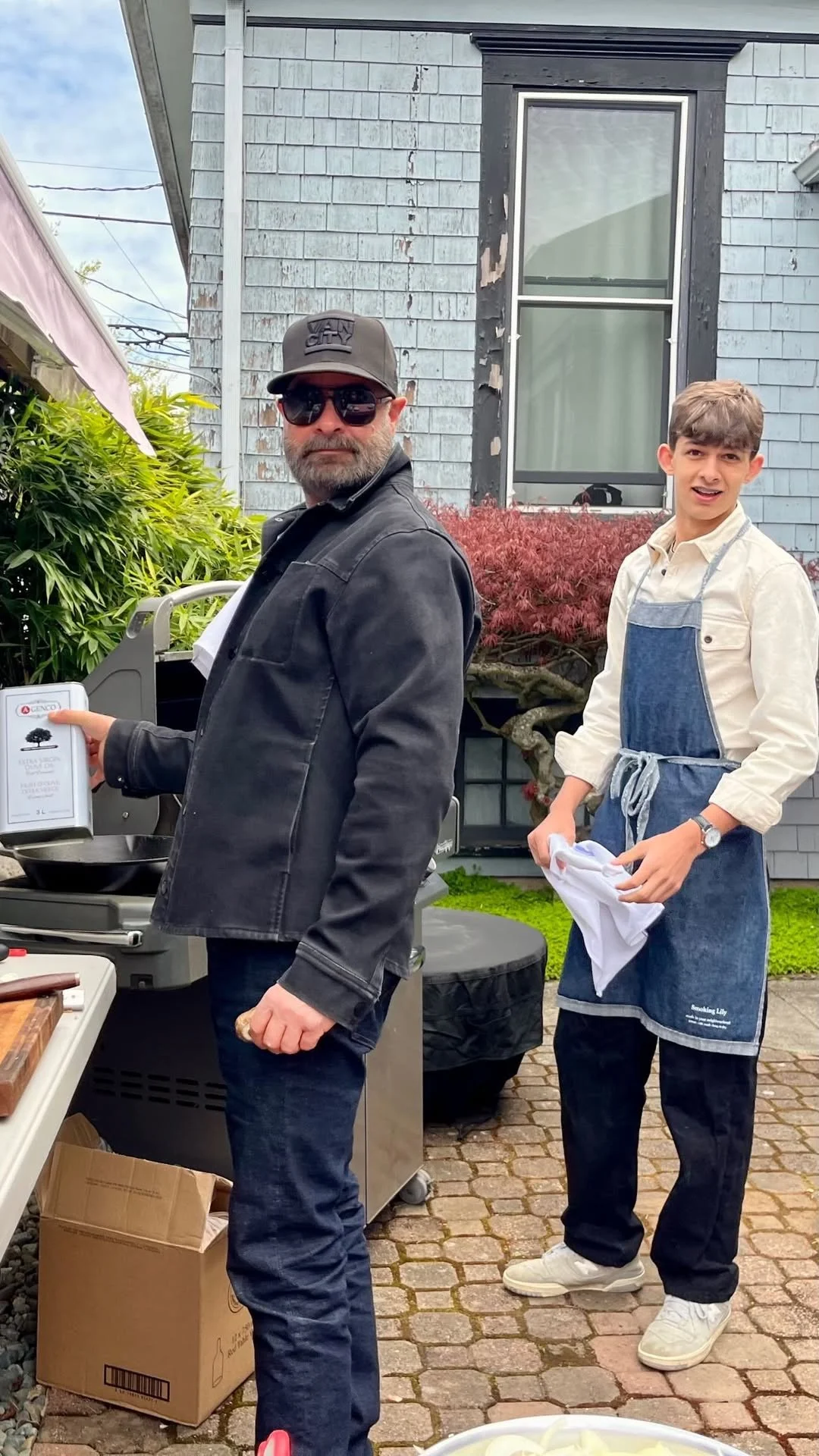 Two men preparing food outdoors in a backyard. The man on the left wears sunglasses, a cap, and a black jacket, holding a bottle of cooking oil. The man on the right is wearing a white shirt, a blue apron, and white shoes, holding a white cloth.