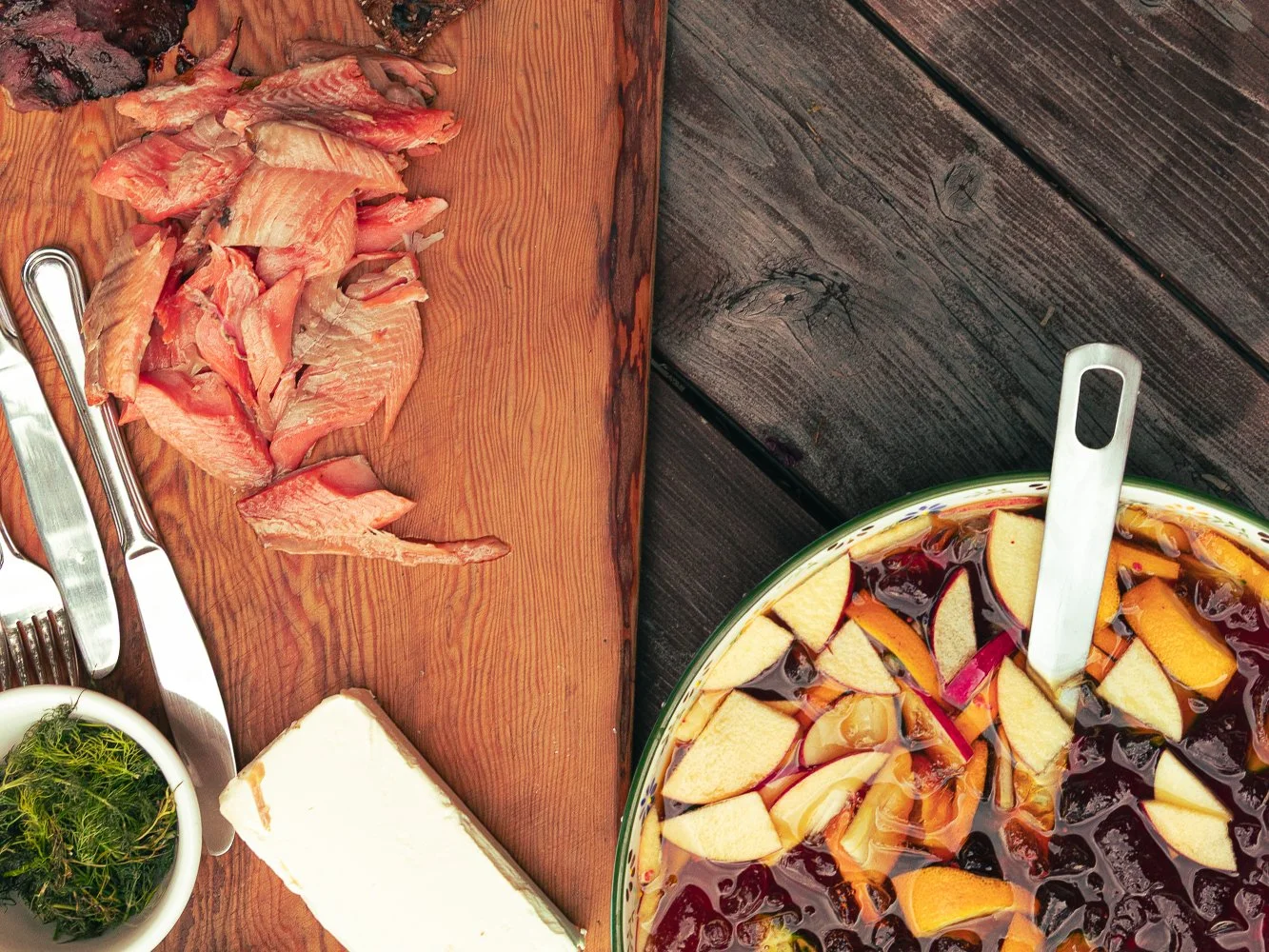 Sliced smoked fish on a wooden cutting board next to a large bowl of fruit salad containing melon, berries, and citrus slices, on a dark wooden table.