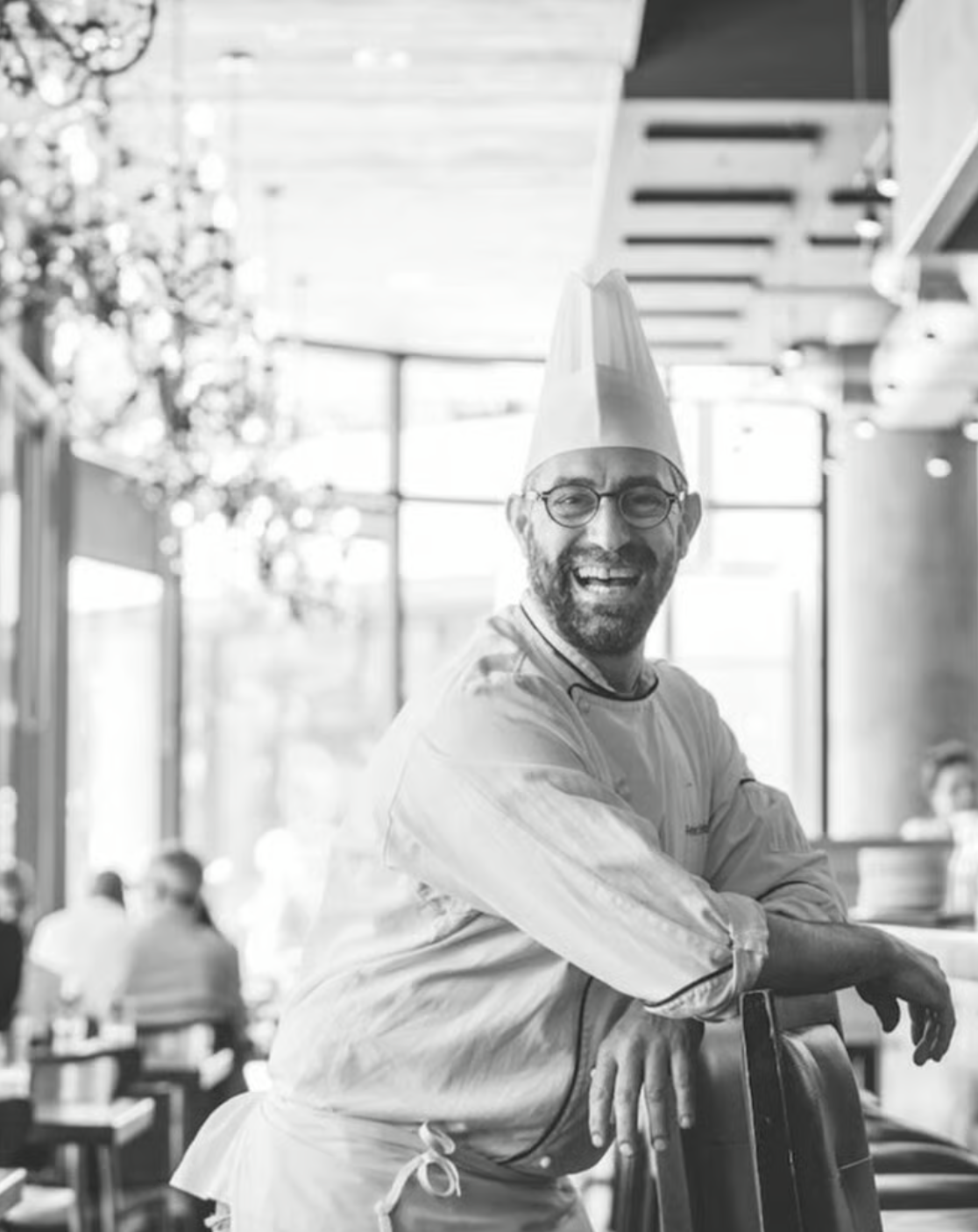 A cheerful chef with glasses and a beard wearing a chef's hat and apron, smiling in a restaurant or cafe with large windows and other people in the background.