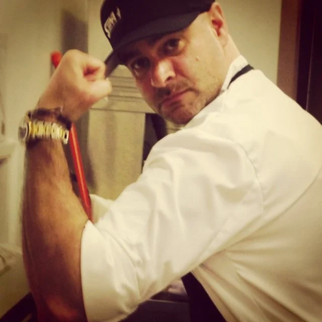 A man in a white shirt and black cap flexing his right arm to show his bicep.