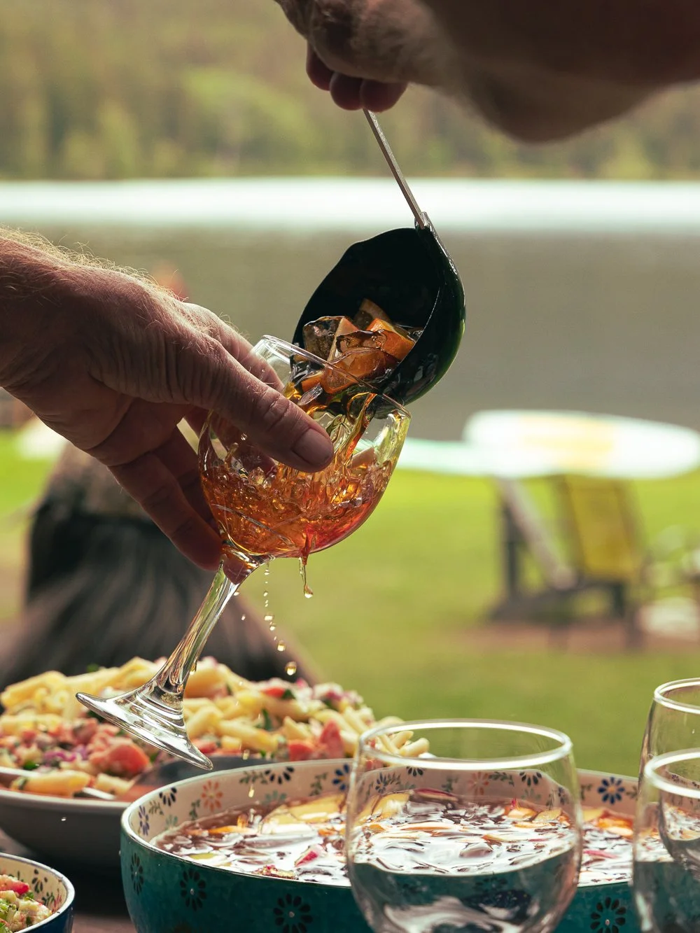 Person pouring dark-colored soda from a bottle into a wine glass filled with ice and beer, with food and other glasses on a table outdoors.