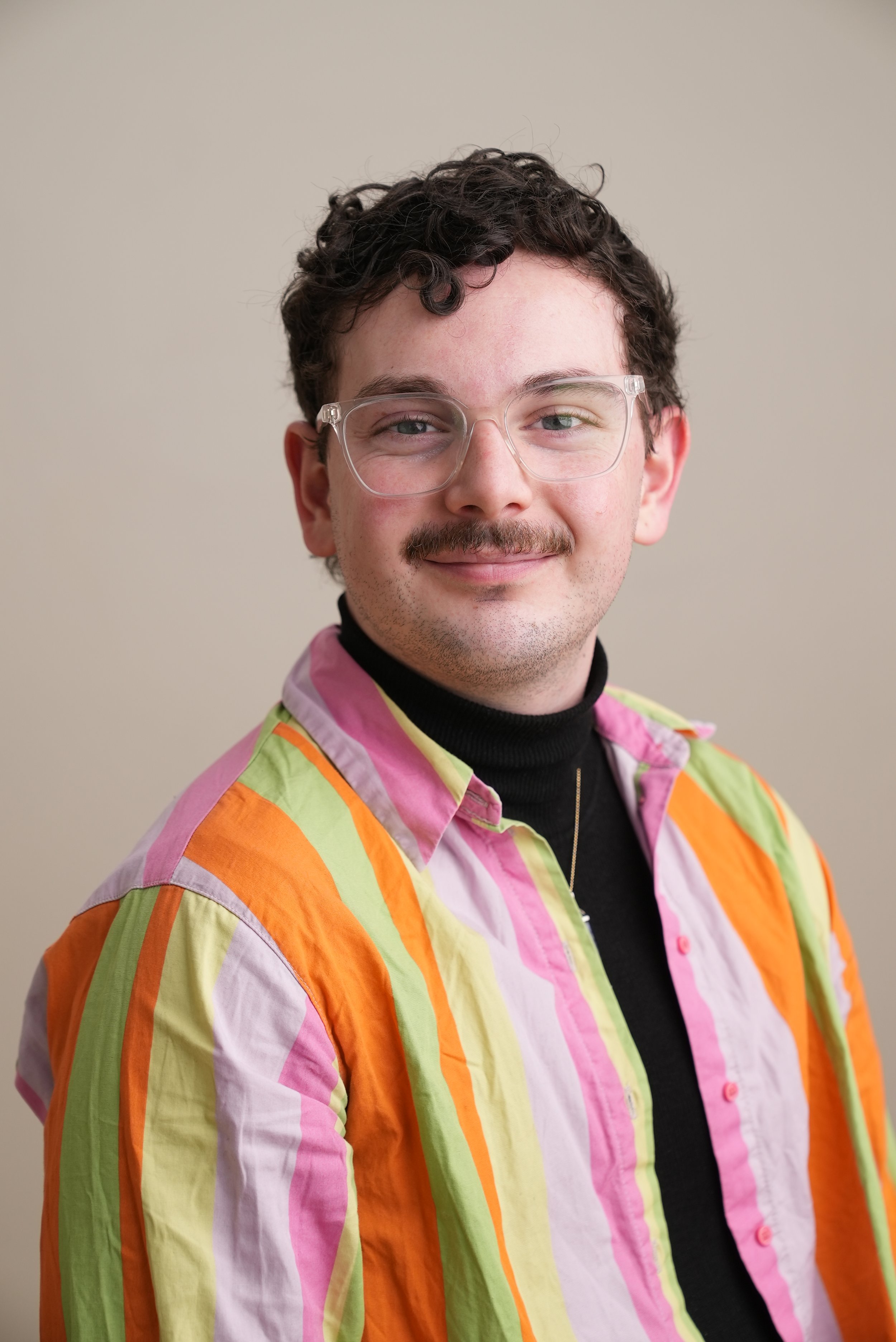 A young man with curly brown hair, wearing clear glasses, smiling, dressed in a black turtleneck and a colorful striped shirt with pink, orange, yellow, and green vertical stripes, against a plain beige background.