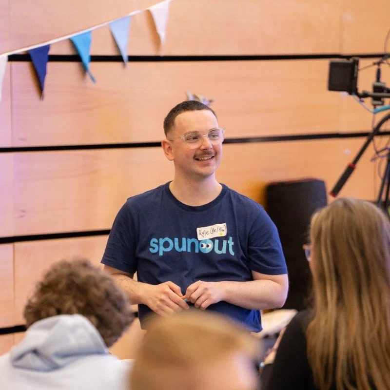 A man smiling and wearing glasses and a blue T-shirt with the word "spunout" standing in front of a group of people in a room decorated with blue and white banners.