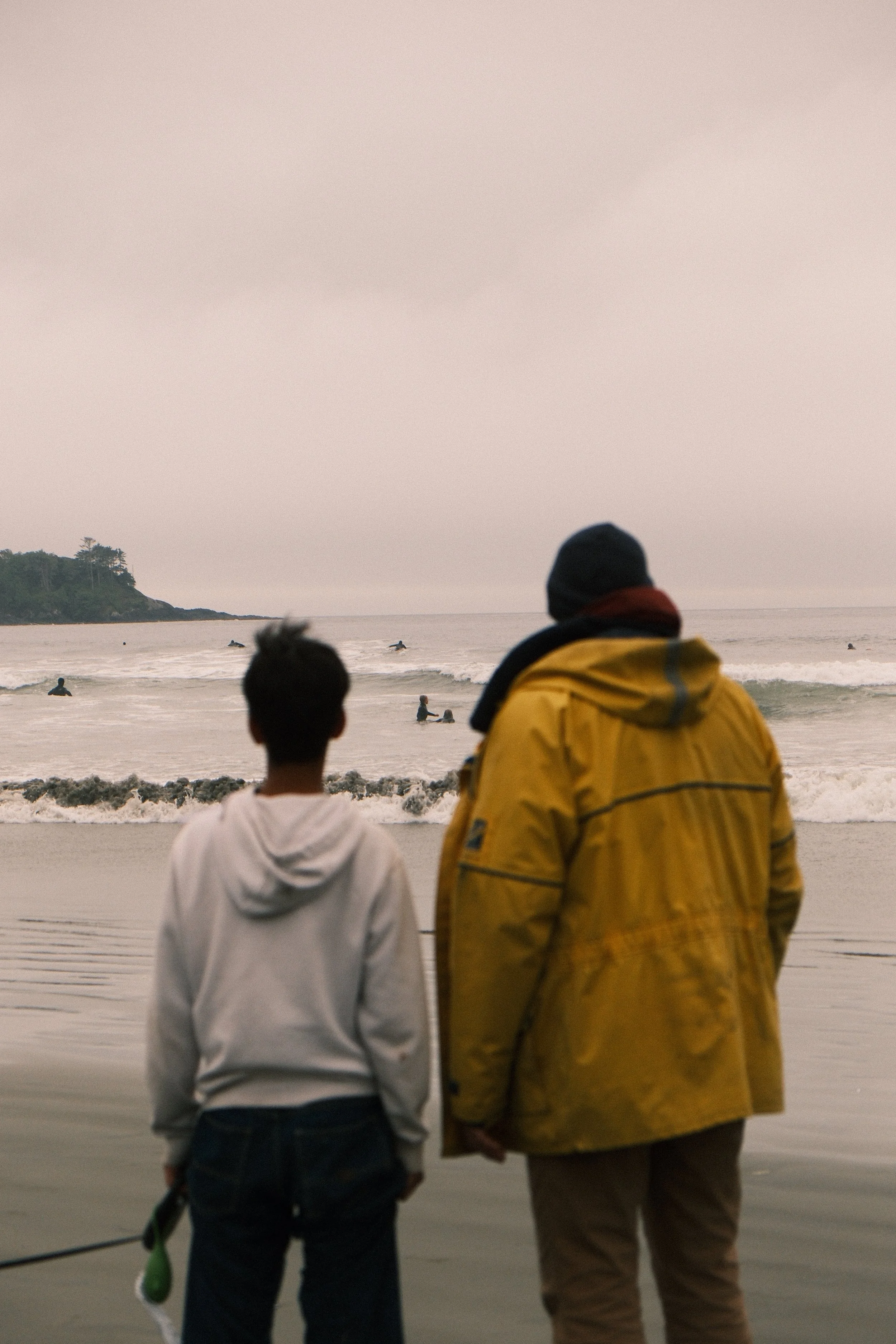 Two people, a child and an adult, are standing on a beach looking at the ocean where people are swimming and surfing, with a small island with trees in the distance, under an overcast sky.