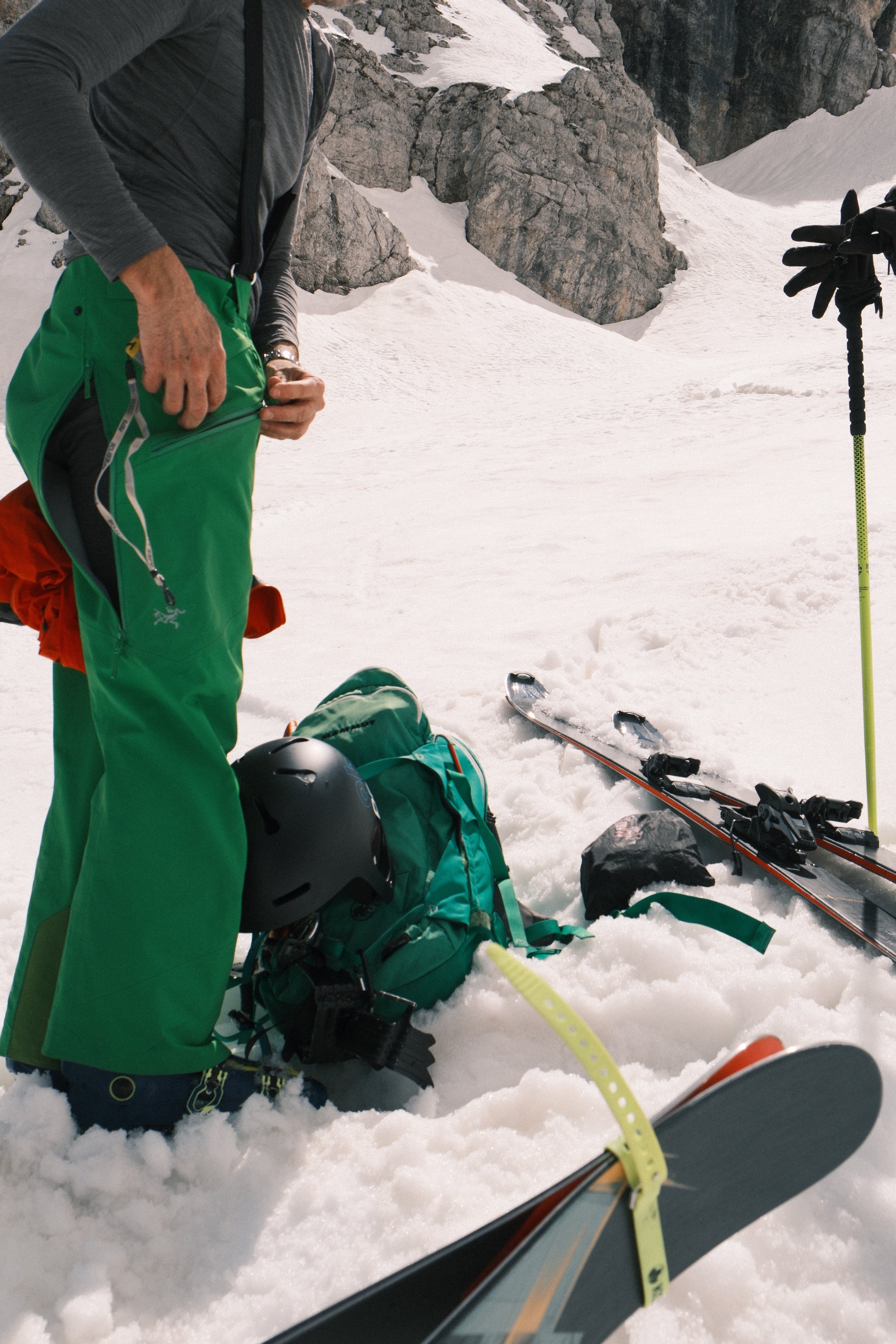 A person in green pants and gray jacket on snowy mountain terrain with climbing gear, skis, and helmets nearby, and rocky cliffs in the background.