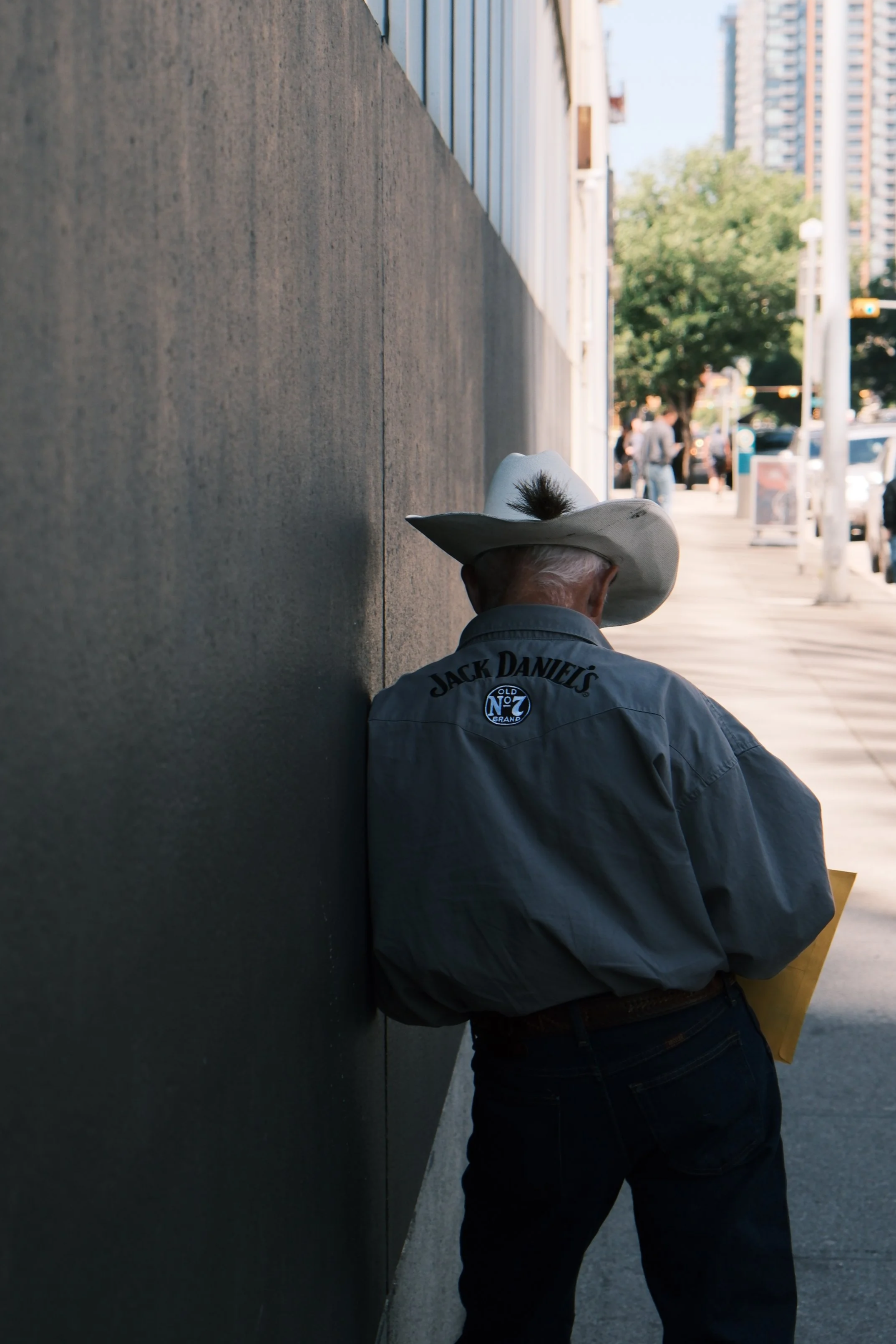 A man in a cowboy hat with a feather, wearing a gray Jack Daniel's shirt, is sitting on the sidewalk beside a large dark wall in an urban area with trees and tall buildings in the background.