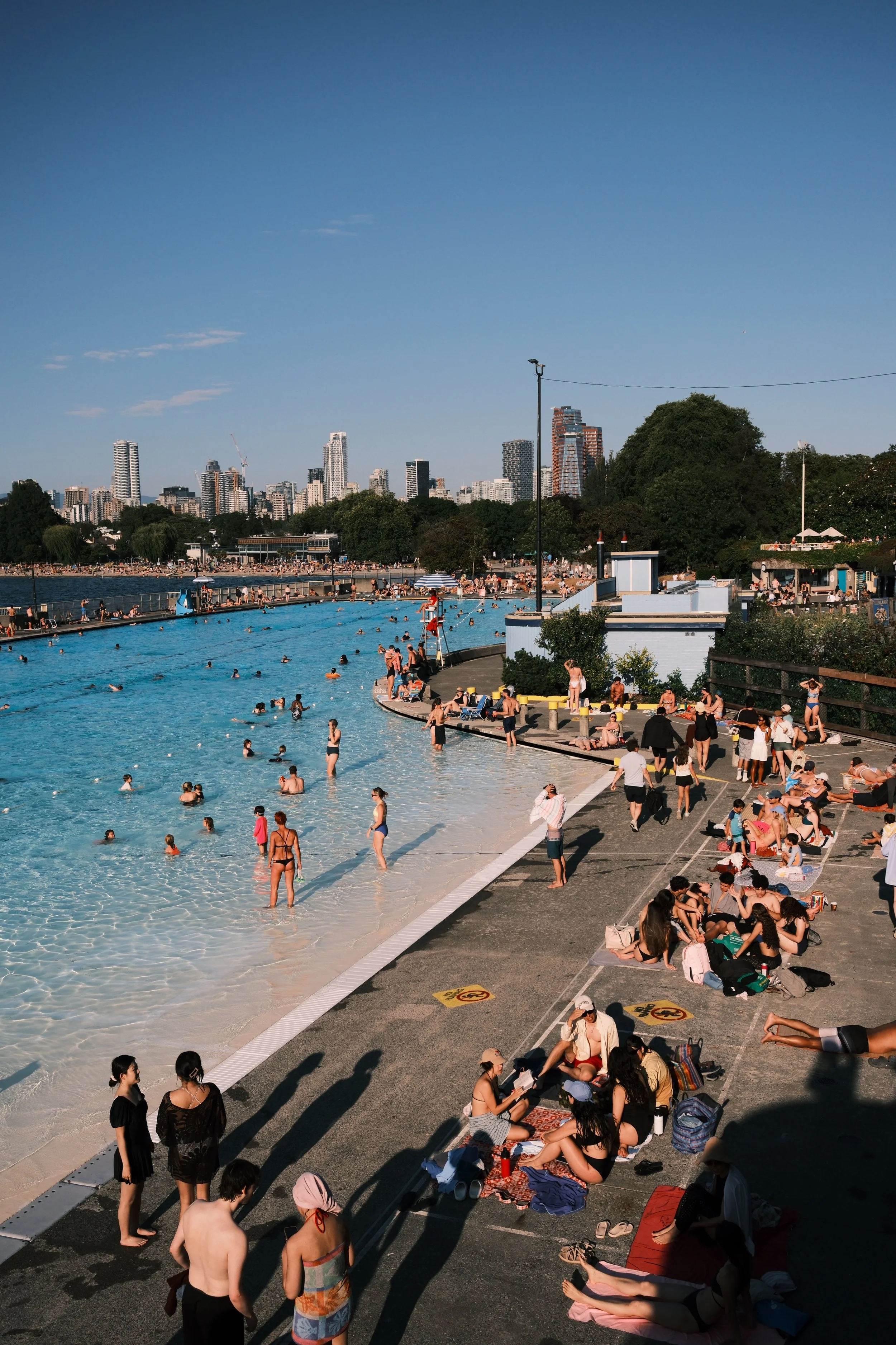People enjoying a day at an outdoor swimming pool near a city skyline with tall buildings, trees, and clear blue sky in the background.