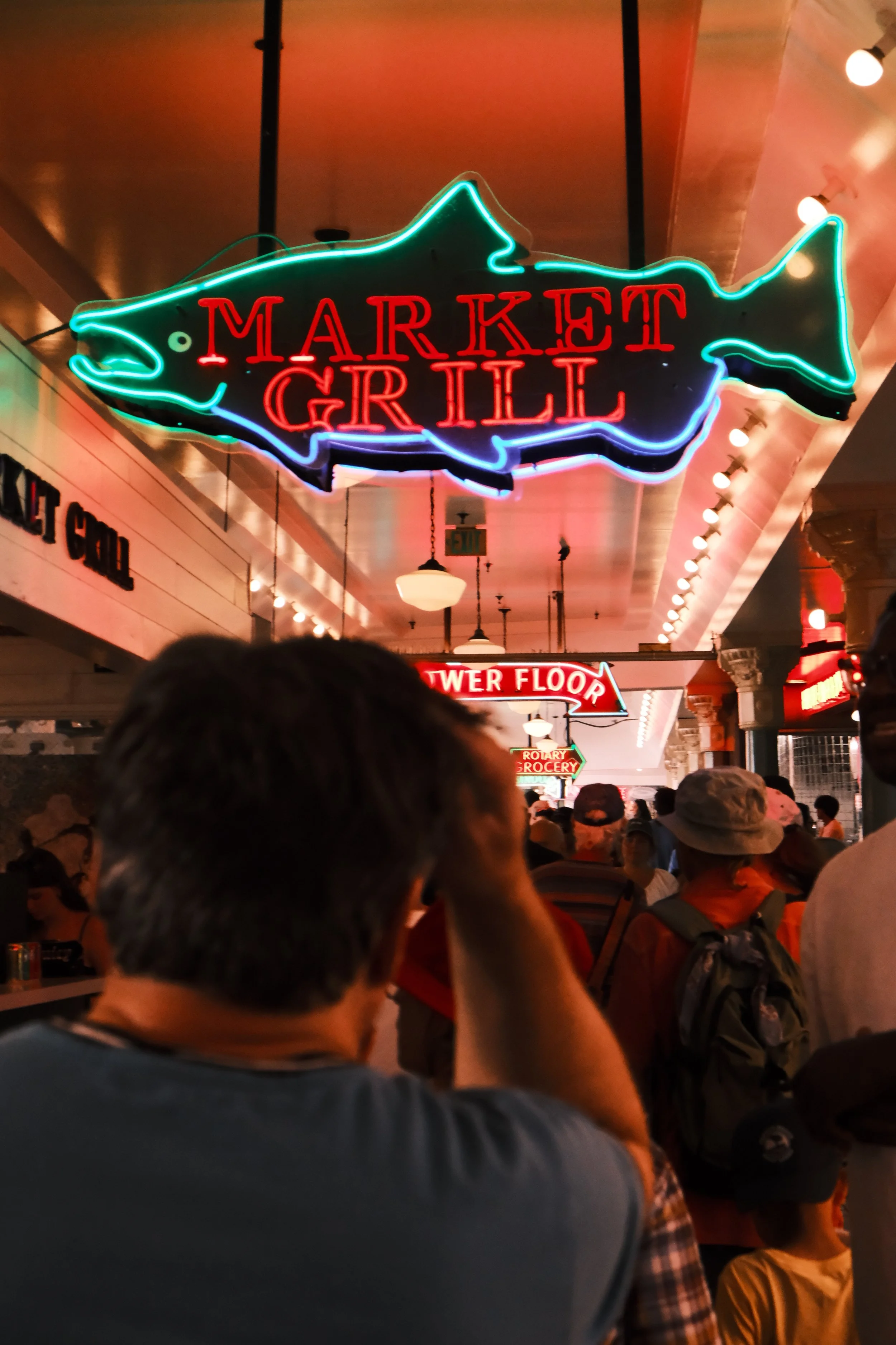Neon sign in the shape of a fish with the words 'Market Grill' inside, hanging in a busy indoor market or food hall with people gathered below.
