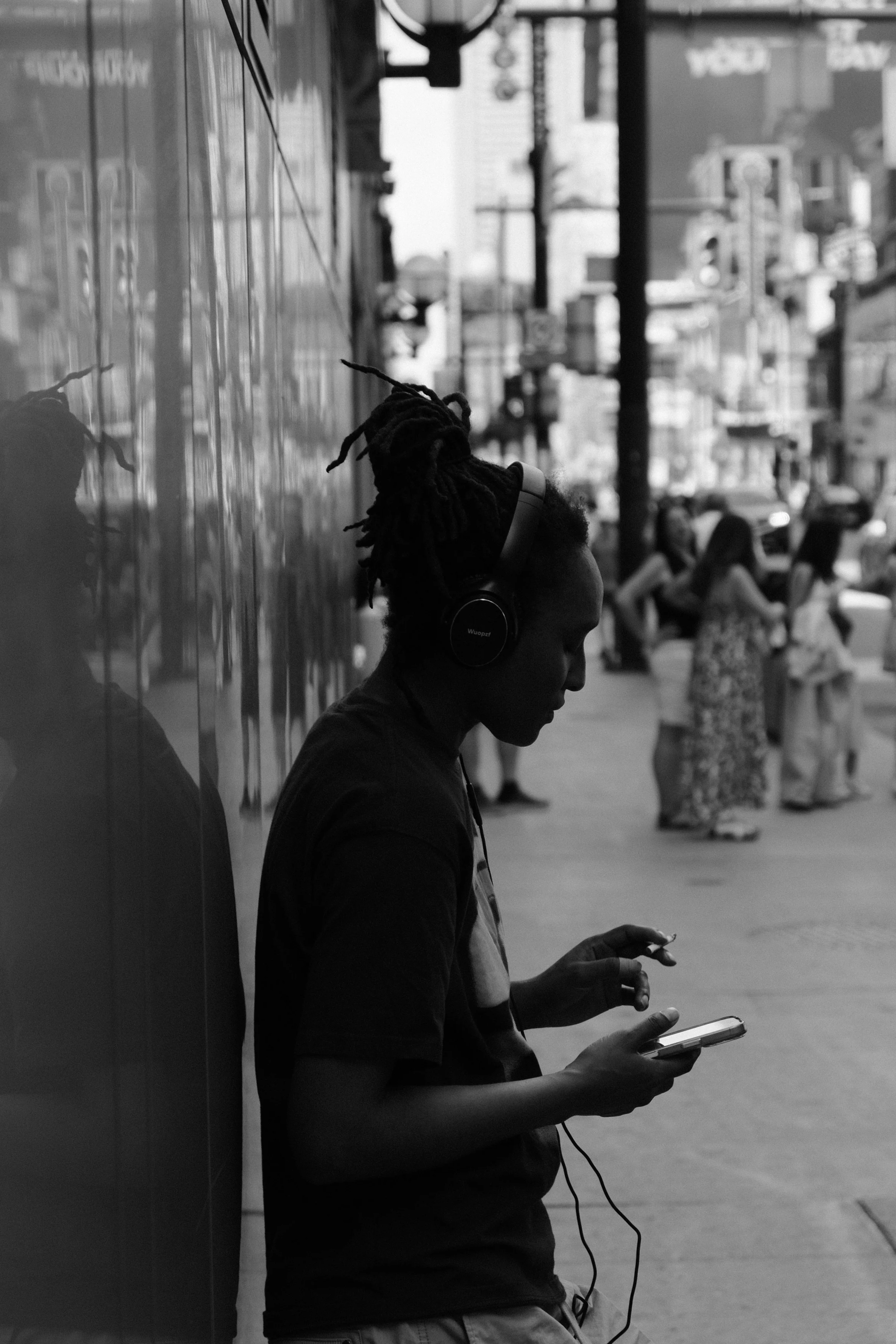 A woman with dreadlocks listening to music through headphones while looking at her phone, leaning against a reflective surface on a city sidewalk with blurred pedestrians in the background.