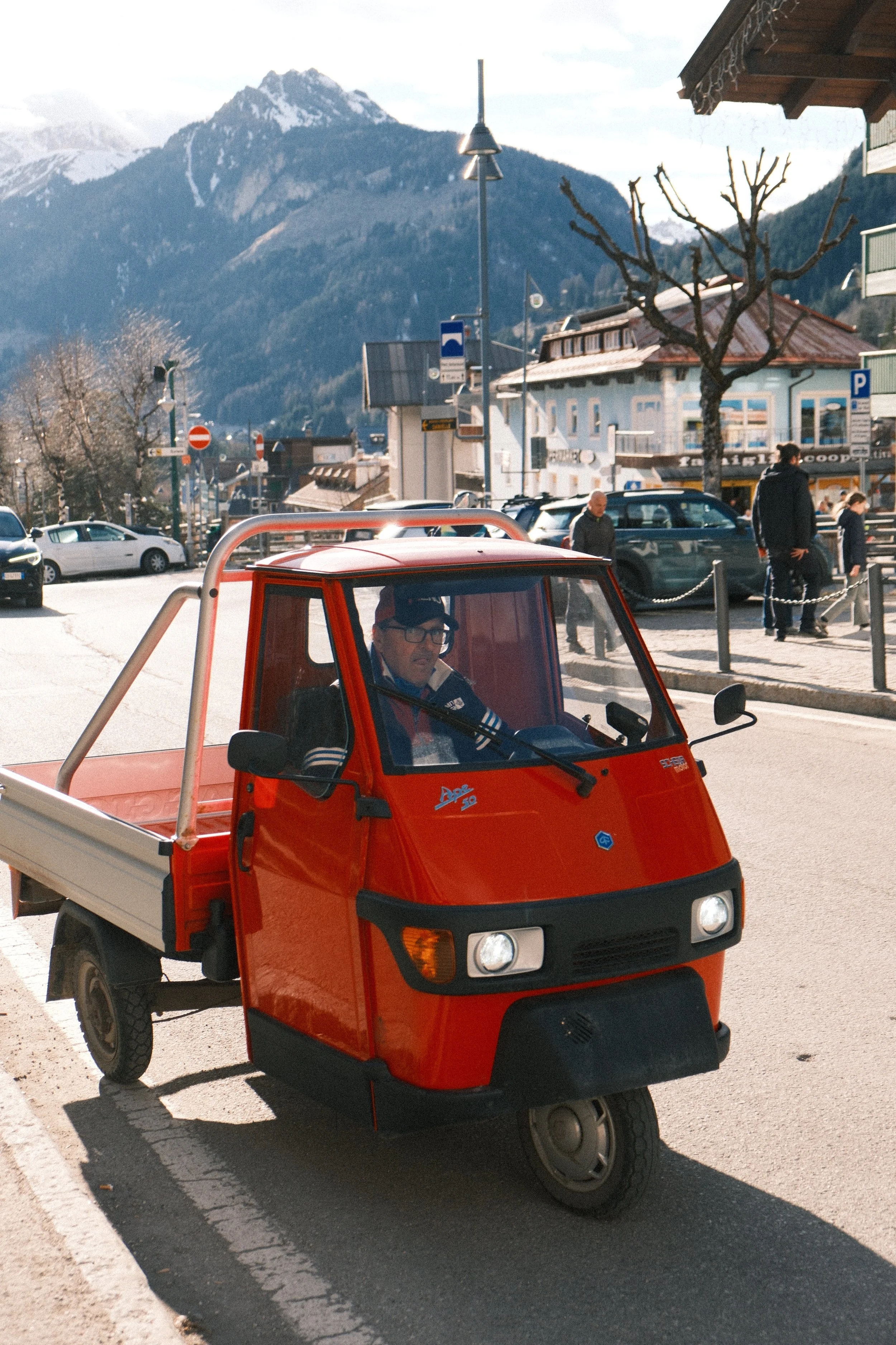 A red three-wheeled utility vehicle with a driver wearing glasses and a dark jacket, parked in a busy town square with mountains in the background. People are walking nearby, and there are buildings, trees, and parked cars.