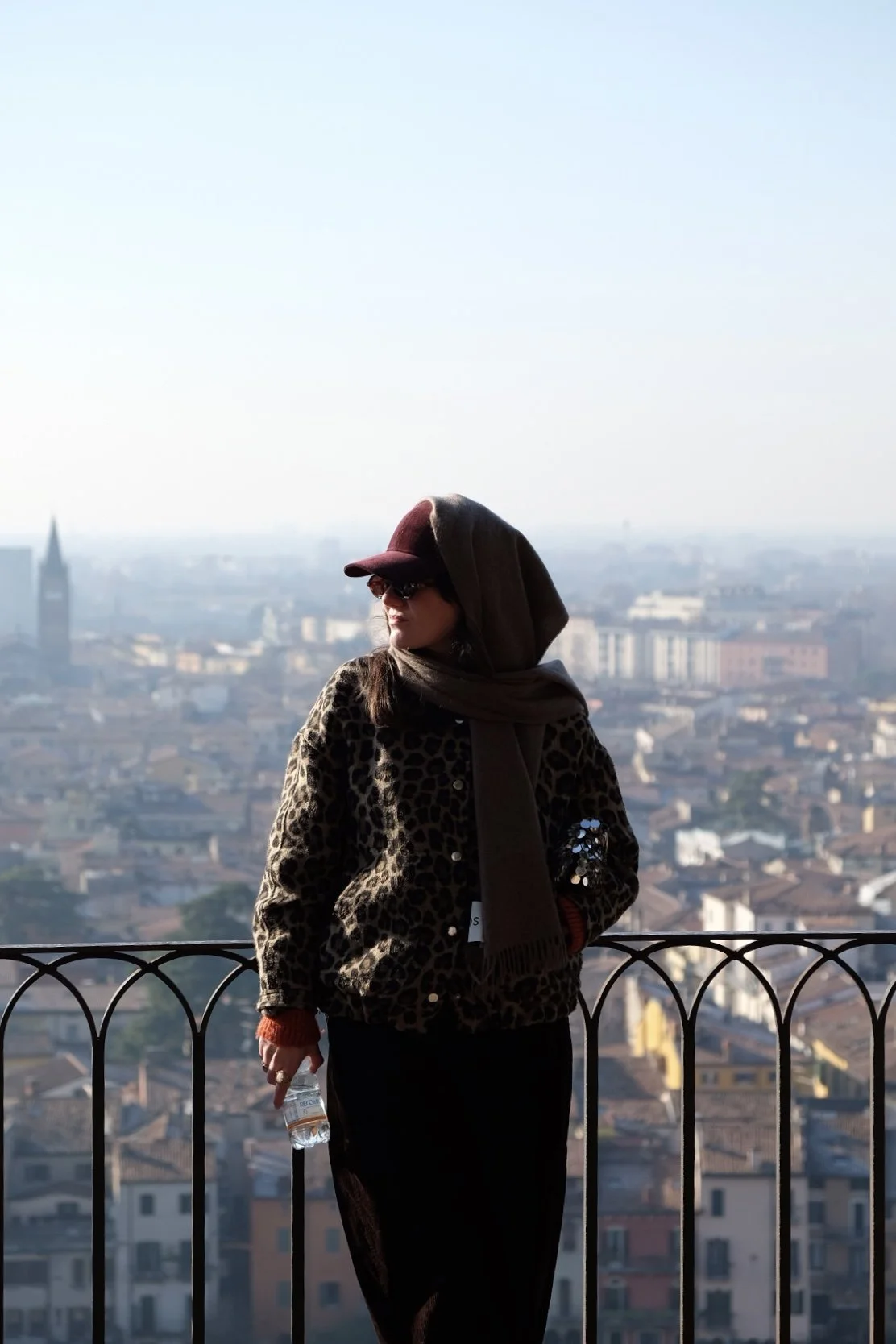 A woman standing on a balcony with a cityscape in the background. She is wearing a leopard print jacket, a dark hoodie, a cap, and sunglasses, holding a water bottle.