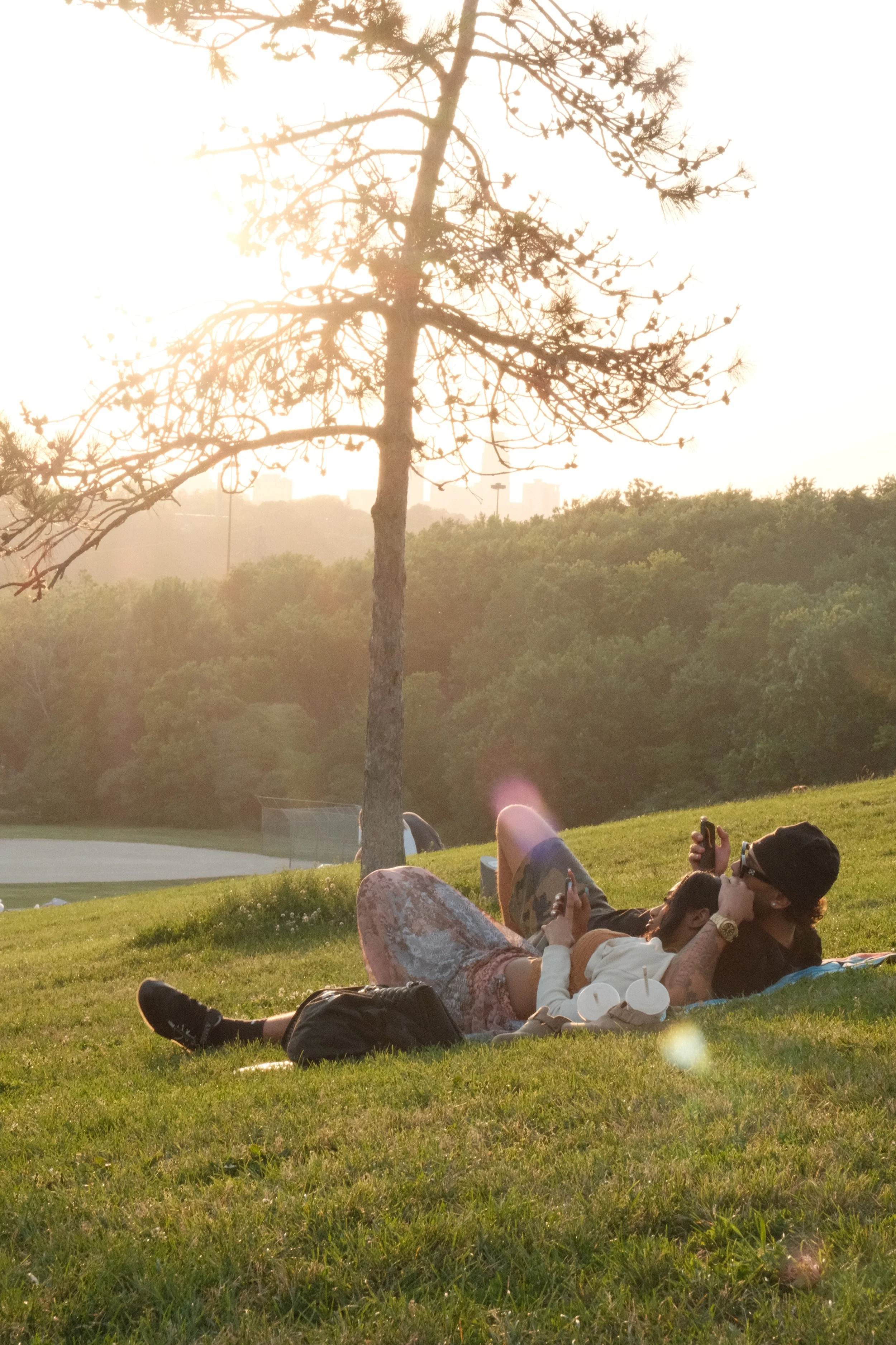 Two people lying on a grassy hillside, relaxing and taking selfies during sunset with trees and distant city buildings in the background.