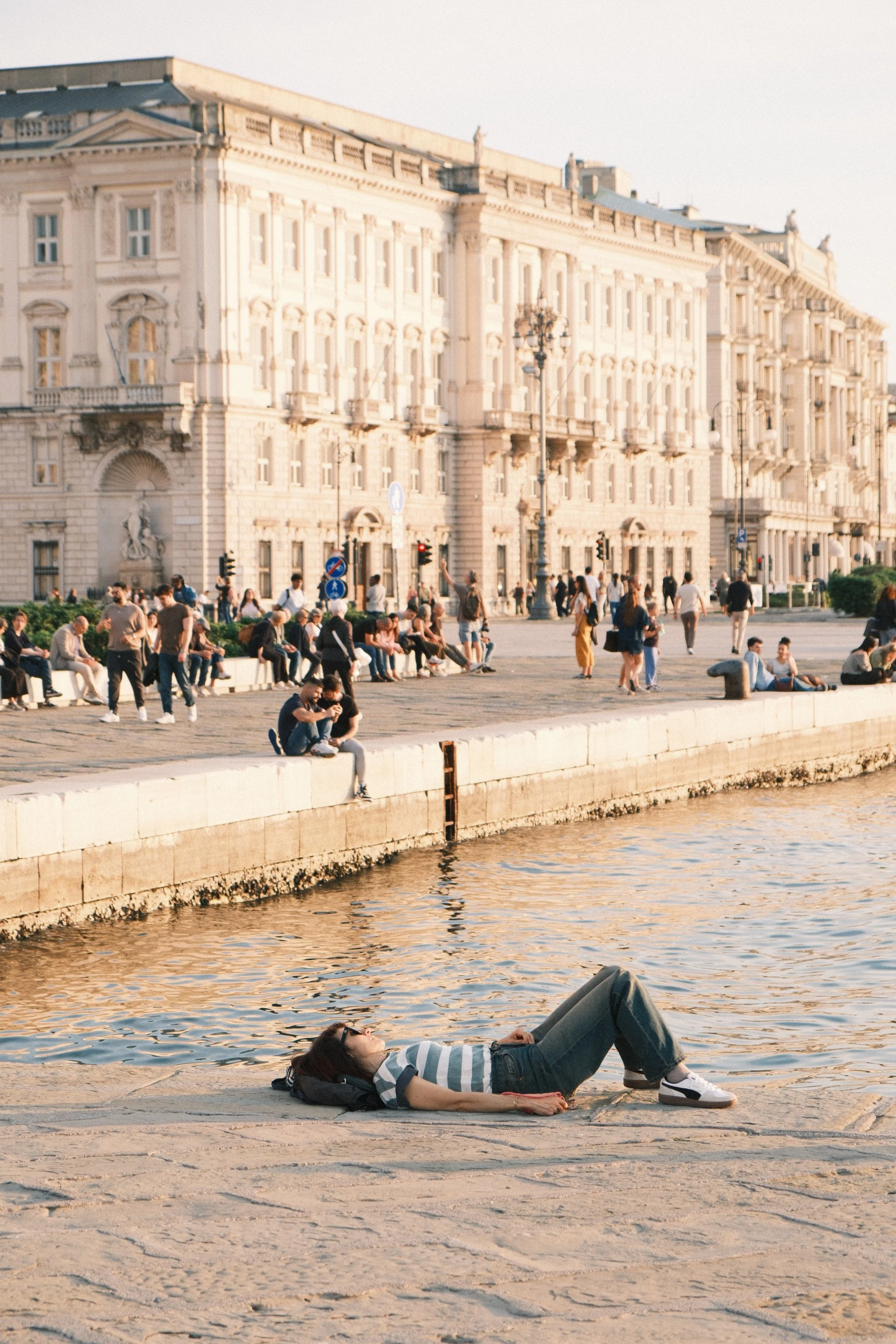 A woman lying on the ground by a waterfront, wearing a striped shirt, jeans, and sneakers, with her eyes closed and a backpack nearby. In the background, there are people walking and sitting along the promenade in front of a large, ornate historic building, with a lamppost and street signs visible.