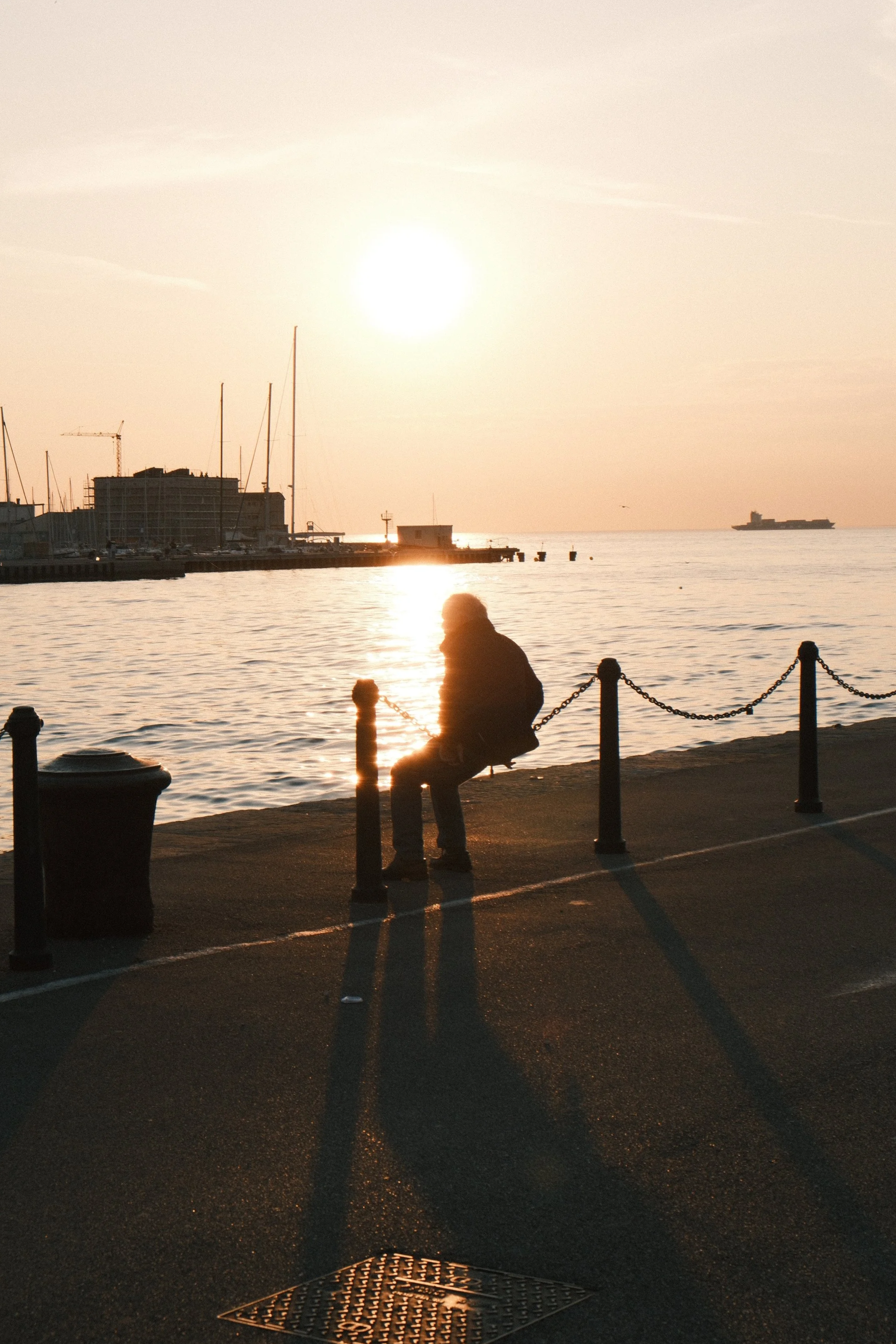 A person sitting alone on a bench by the water during sunset, with the sun low on the horizon, casting a warm glow and long shadows. Several boats are docked nearby, and a cruise ship is visible in the distance. The scene is peaceful and serene.