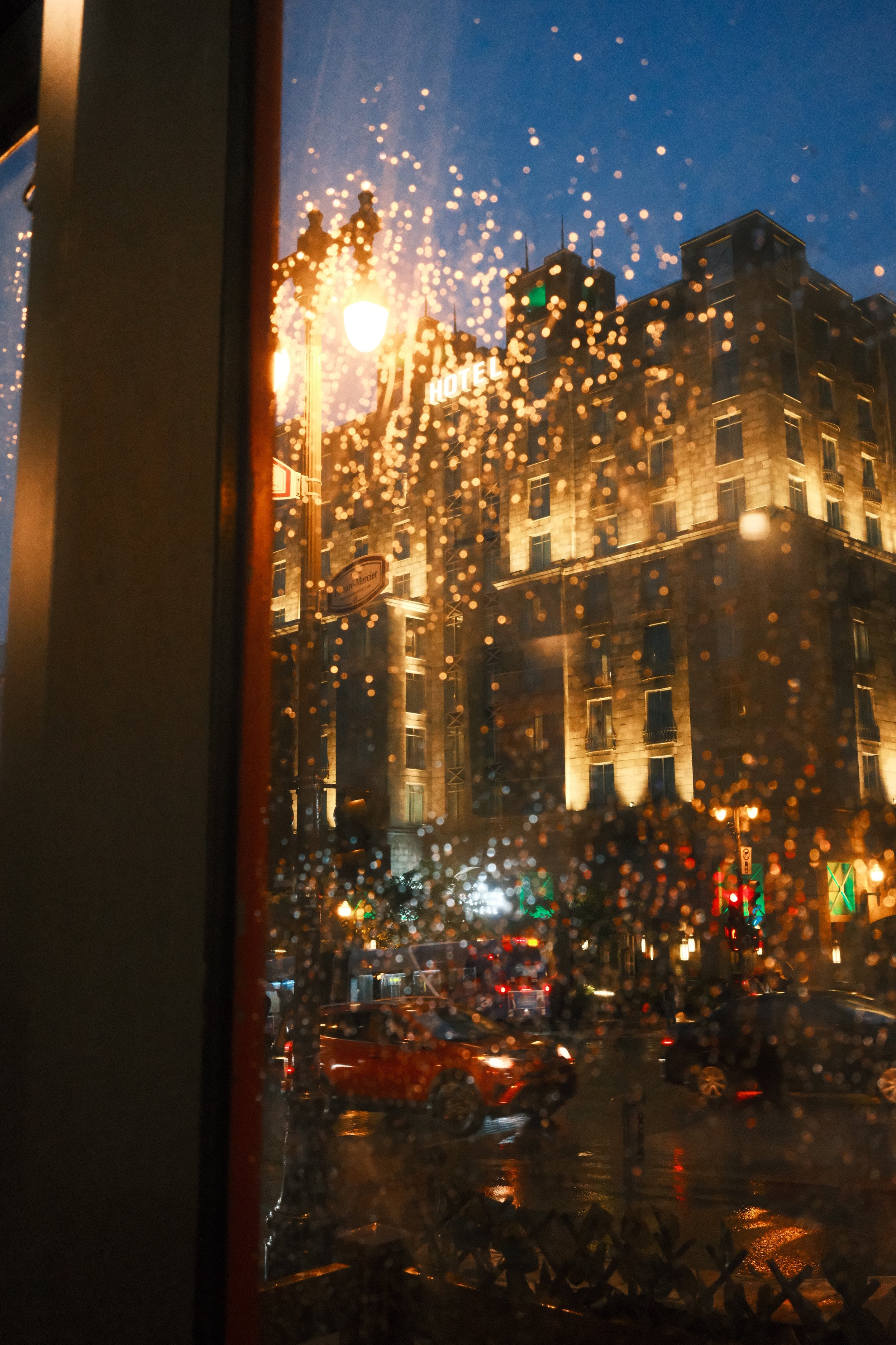 Urban street scene viewed through a rain-speckled window at night. Illuminated building with the word 'HOTEL' visible, street lights, and traffic with car lights reflecting on the wet road.