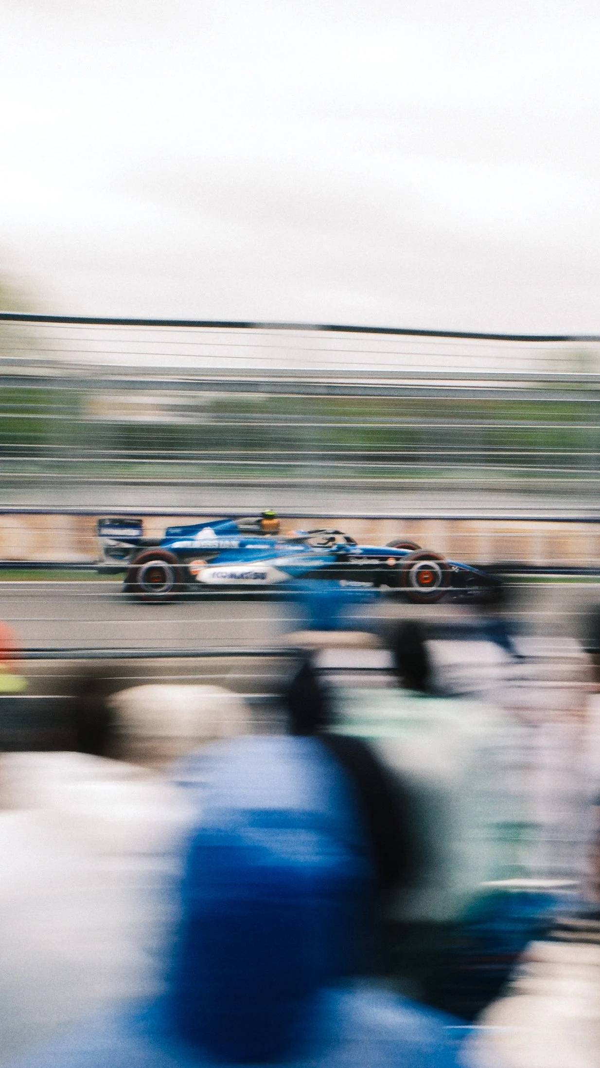 Blurred image of a blue Formula 1 race car speeding on a track with a crowd of spectators in the foreground.