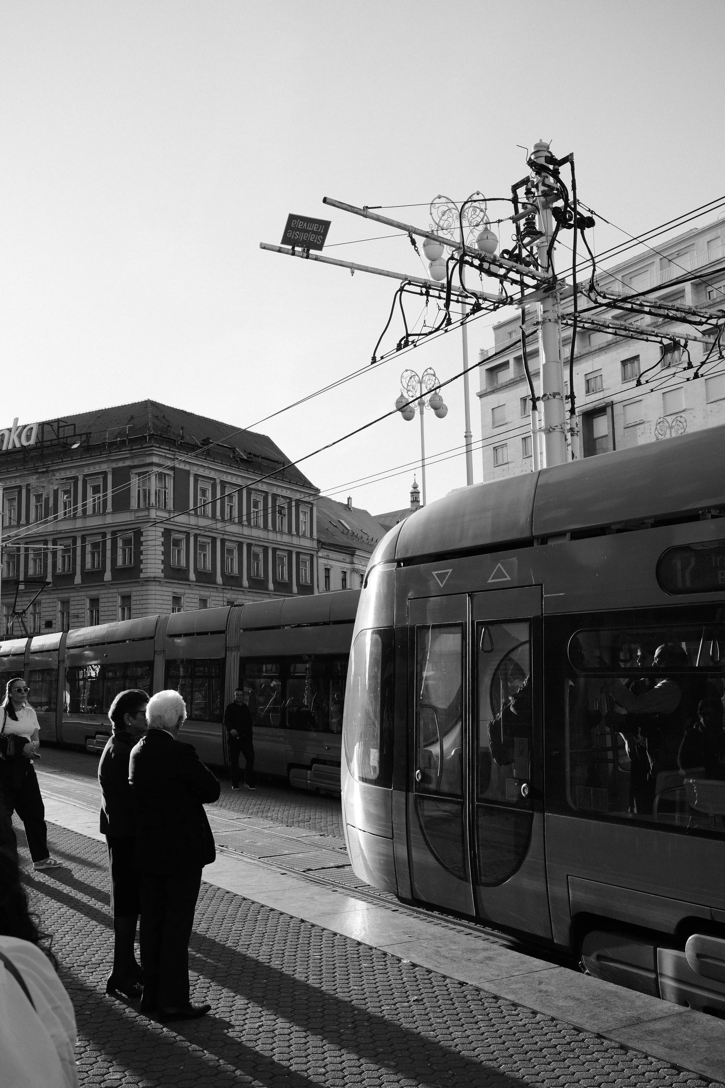 A city street with a modern tram, several pedestrians waiting, and old buildings in the background. Power lines and street lamps are visible.
