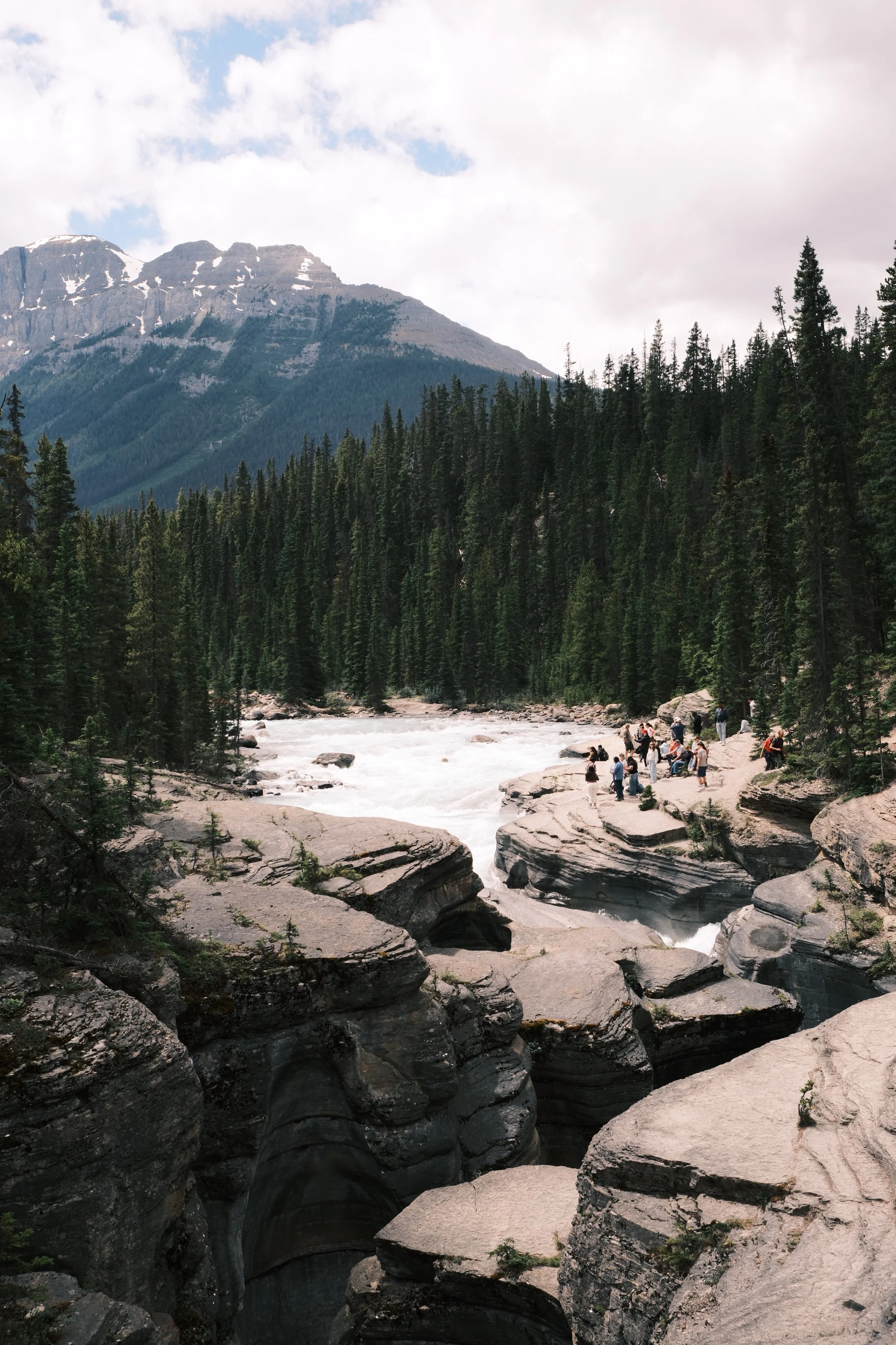 A mountain landscape with snow patches on the peaks, dense evergreen forest, a rushing river, and rocky formations with people exploring near the water.