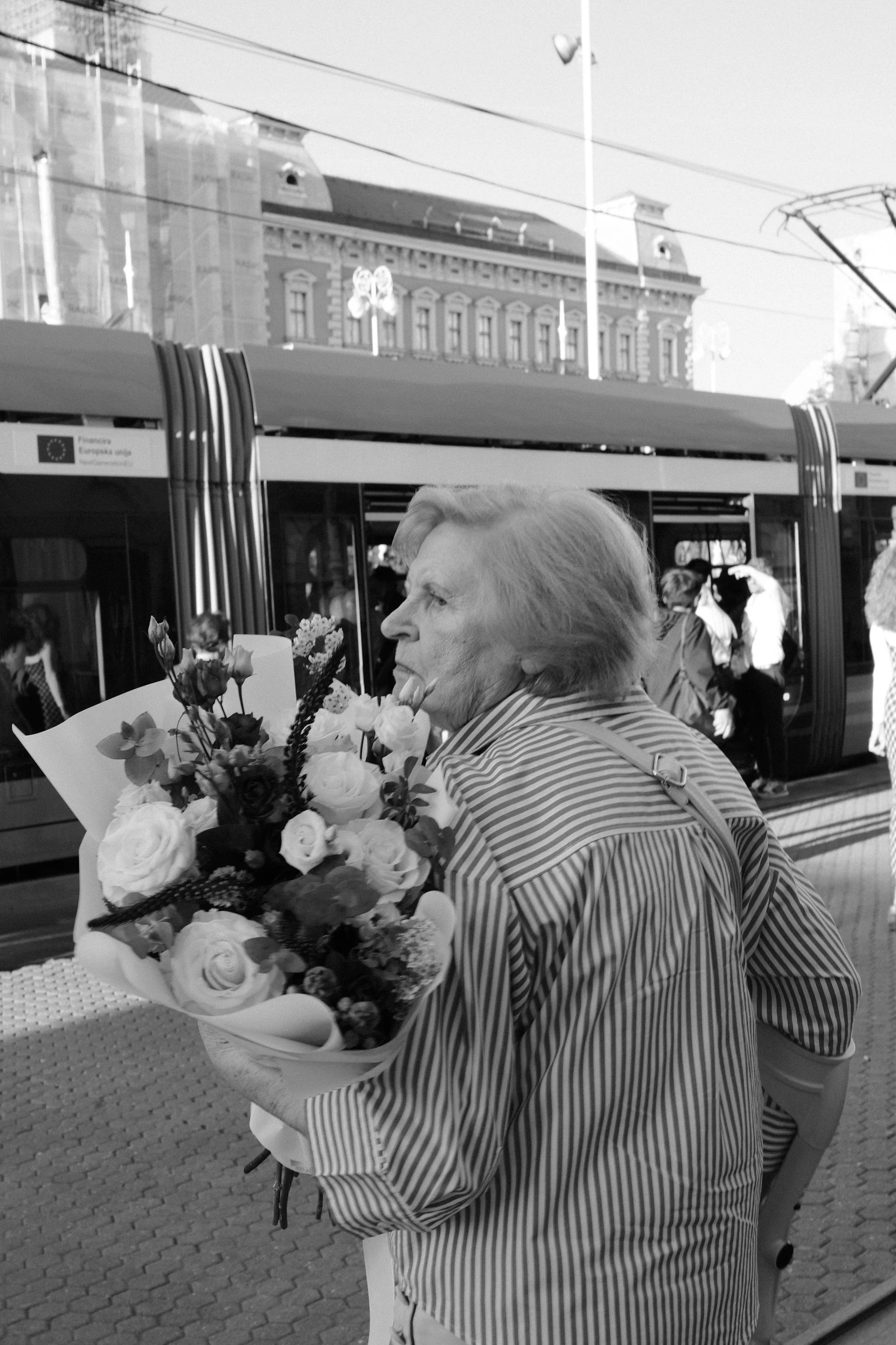 Older woman holding a large bouquet of flowers on a city street, with a city bus and historic buildings in the background.