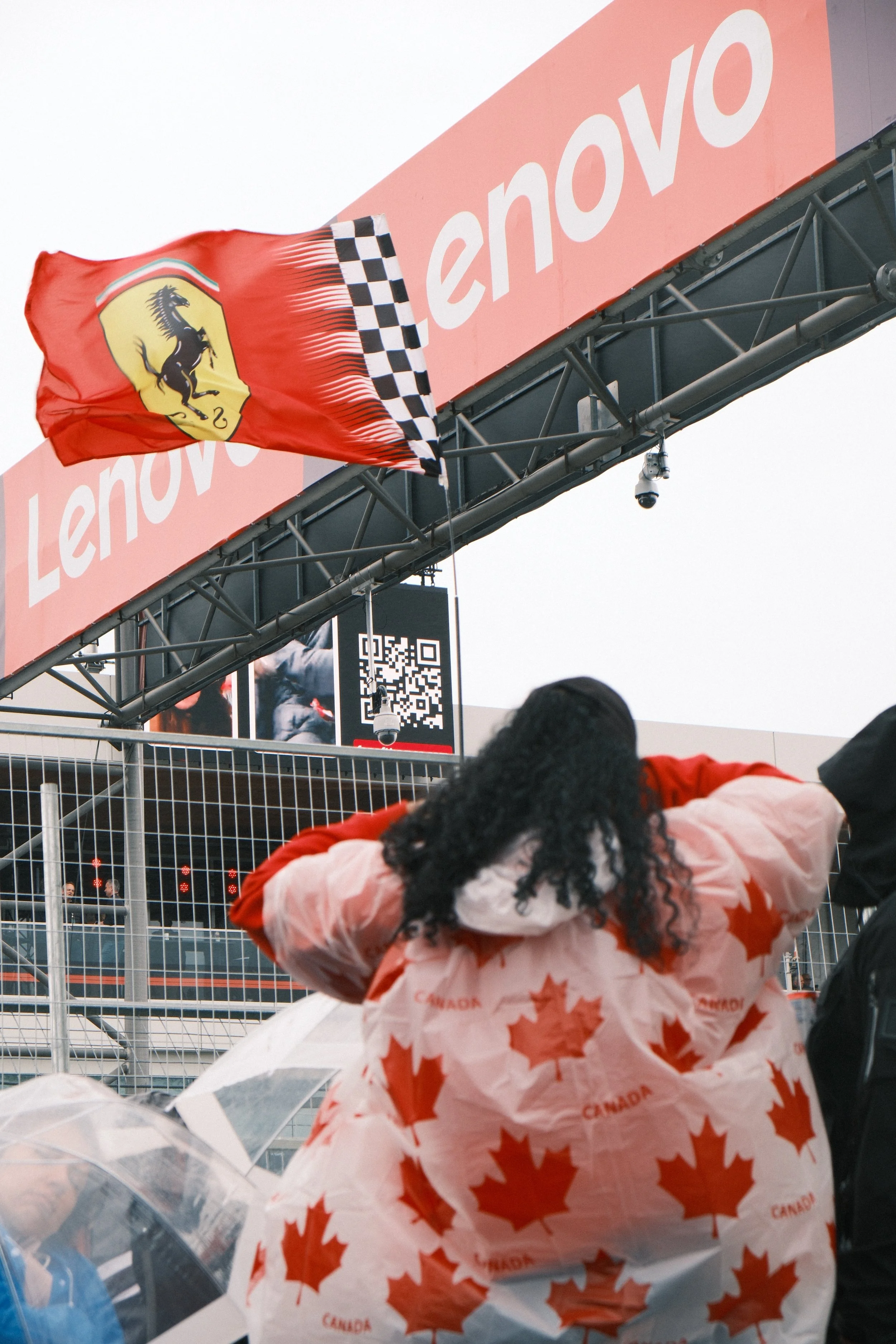People attending a motorsport event, with a woman wearing a Canadian-themed jacket in the foreground and a flag with Ferrari logo and an advertising banner in the background.