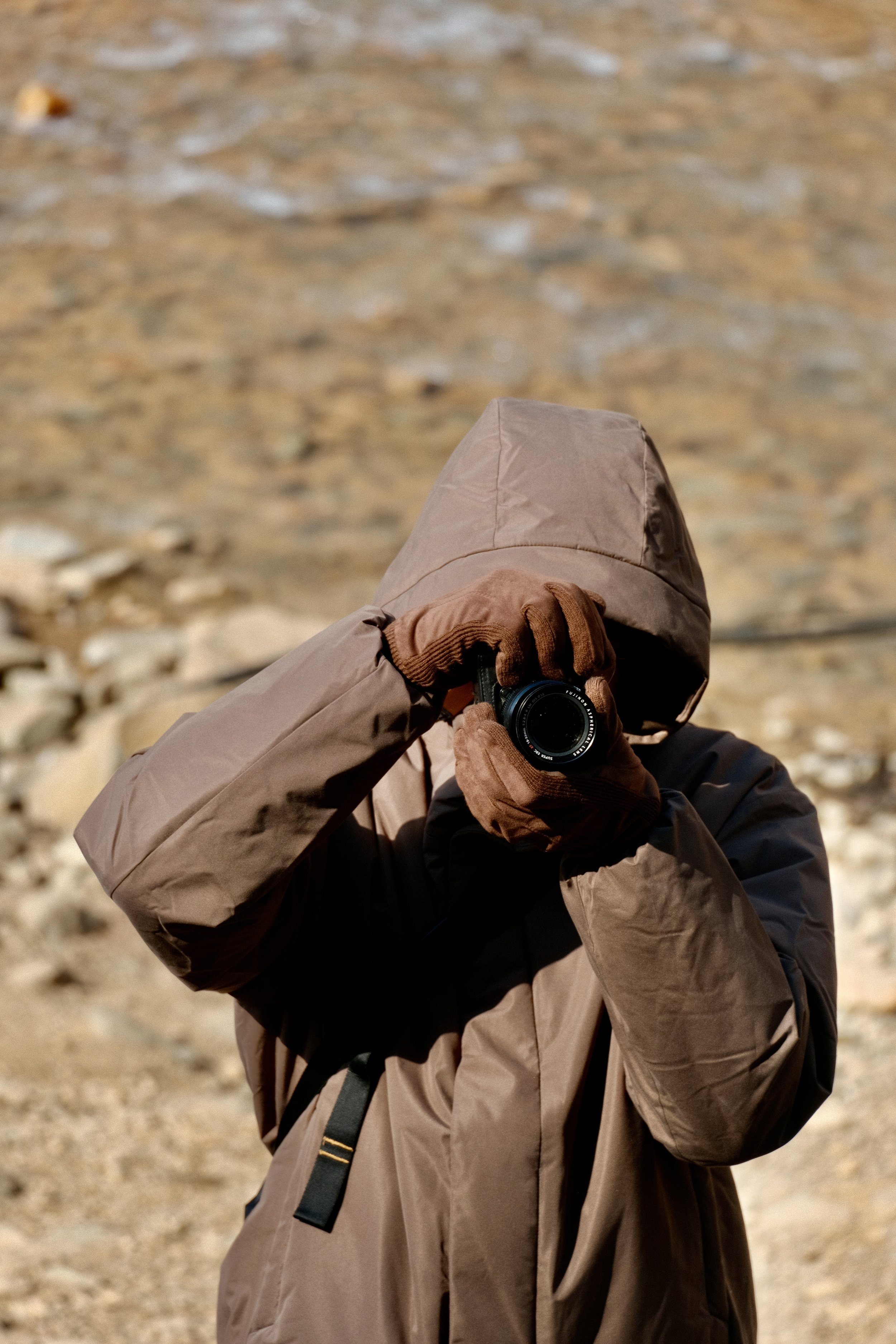 Person dressed in a brown jacket and hood, holding a camera, standing outdoors on rocky terrain.
