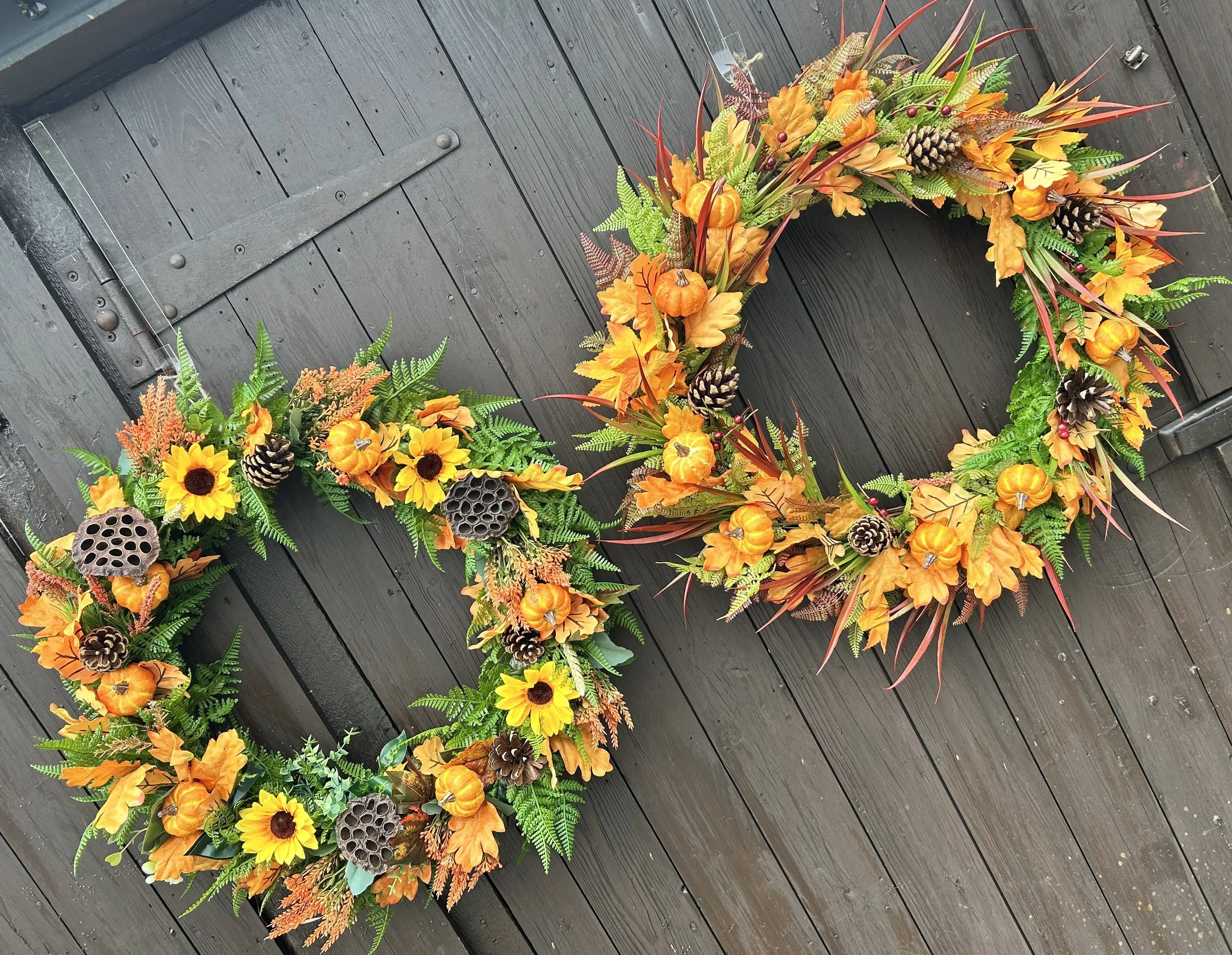 A decorative autumn wreath with orange pumpkins, sunflowers, ferns, pinecones, and various fall leaves, hanging on a dark wooden fence.
