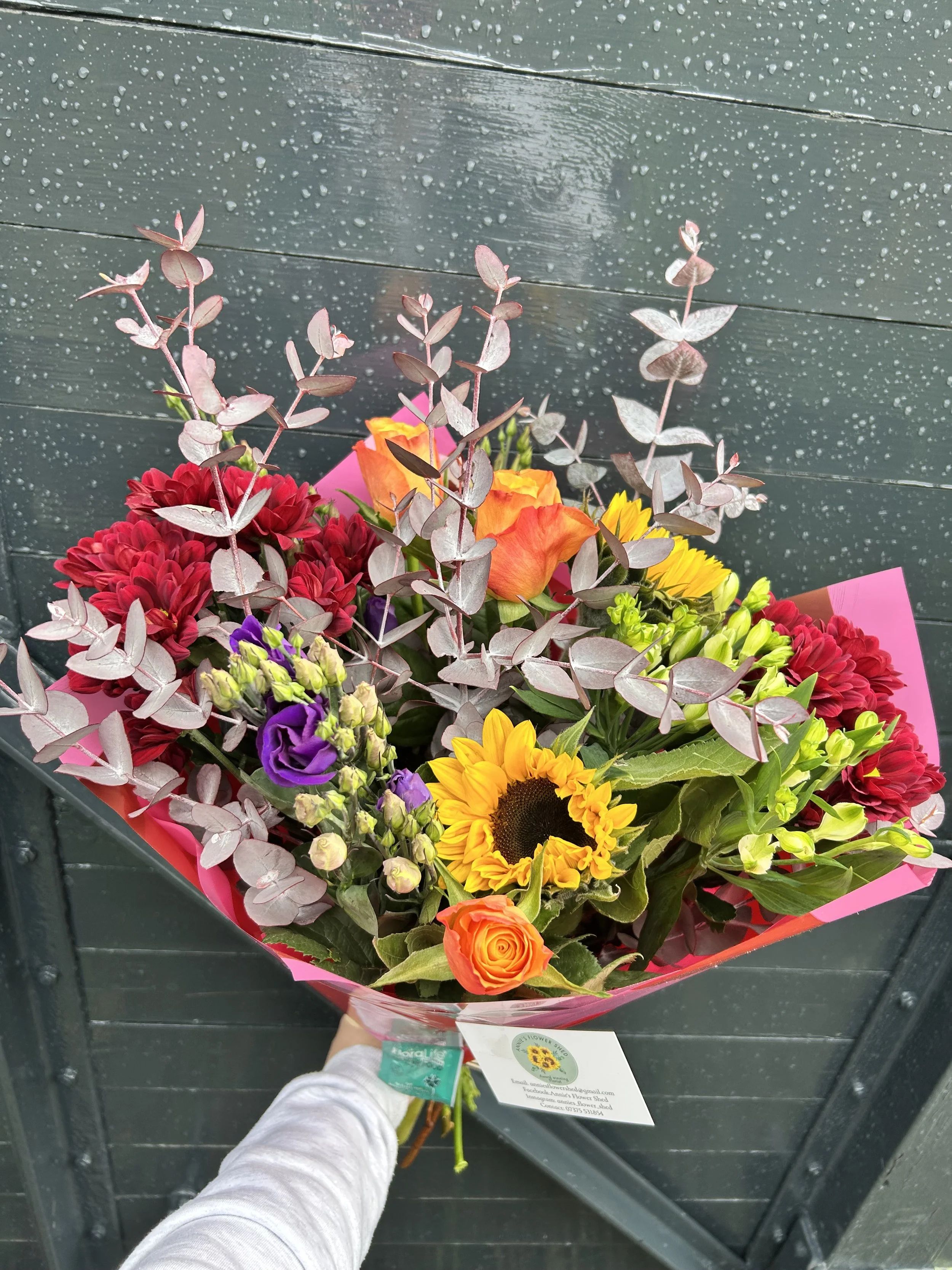 A person holding a colorful bouquet of flowers against a dark, wet, wooden background. The bouquet includes a sunflower, orange and yellow roses, red chrysanthemums, purple lisianthus, and silver dollar eucalyptus leaves, with pink wrapping paper.