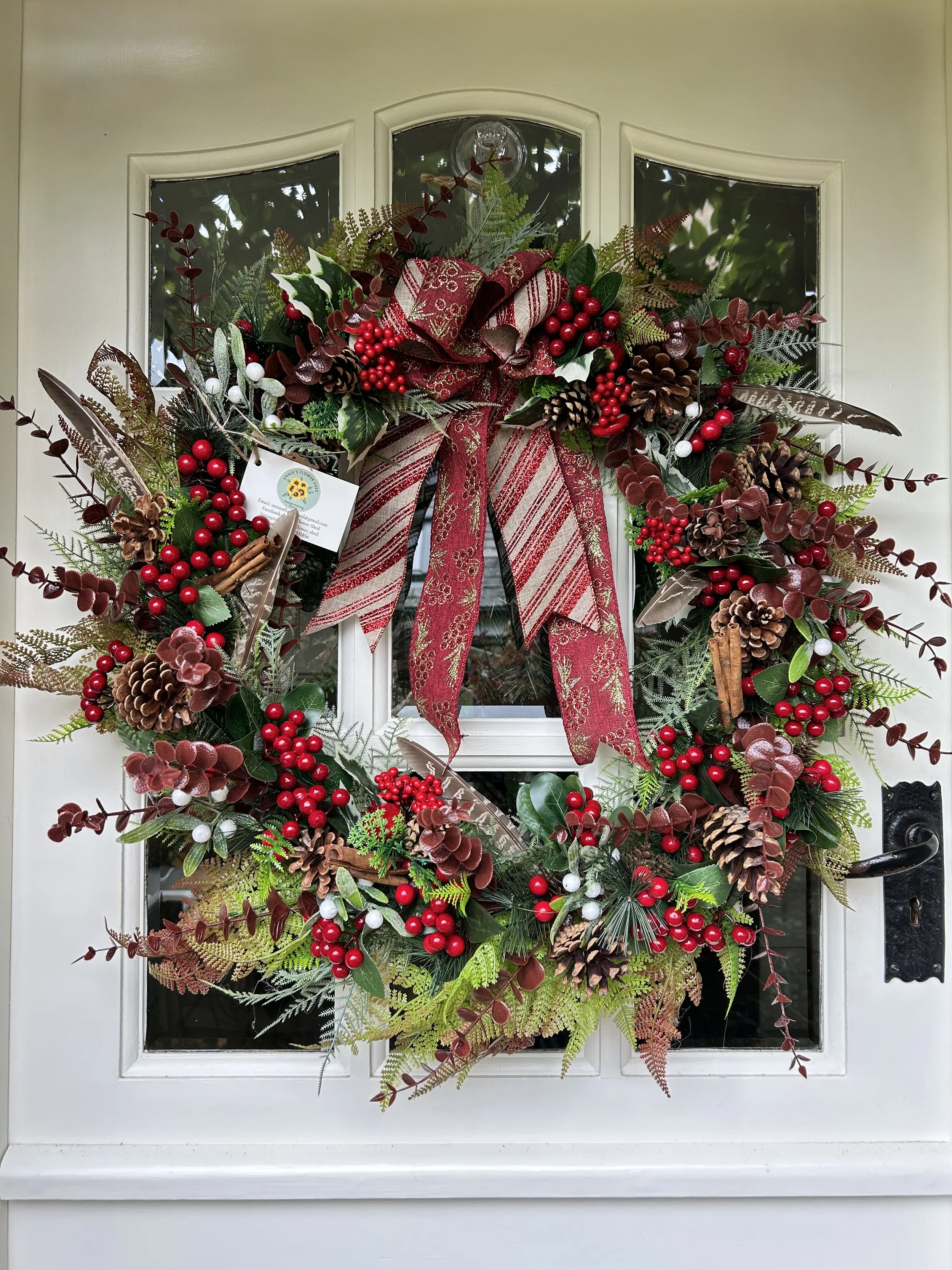 A Christmas wreath decorated with red berries, pinecones, green leaves, and a red and gold ribbon, hanging on a white door with glass panels.