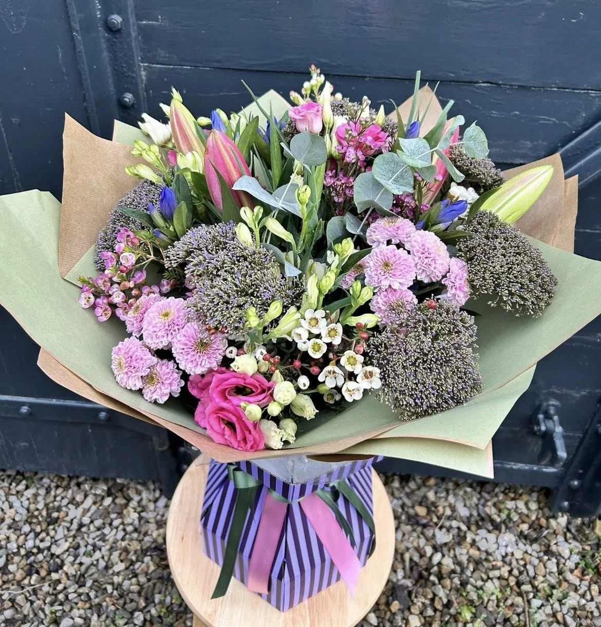 Colorful bouquet of various pink, purple, white lilies, chrysanthemums, and other flowers in striped purple and pink wrapping paper, placed on a wooden stool with a black background.