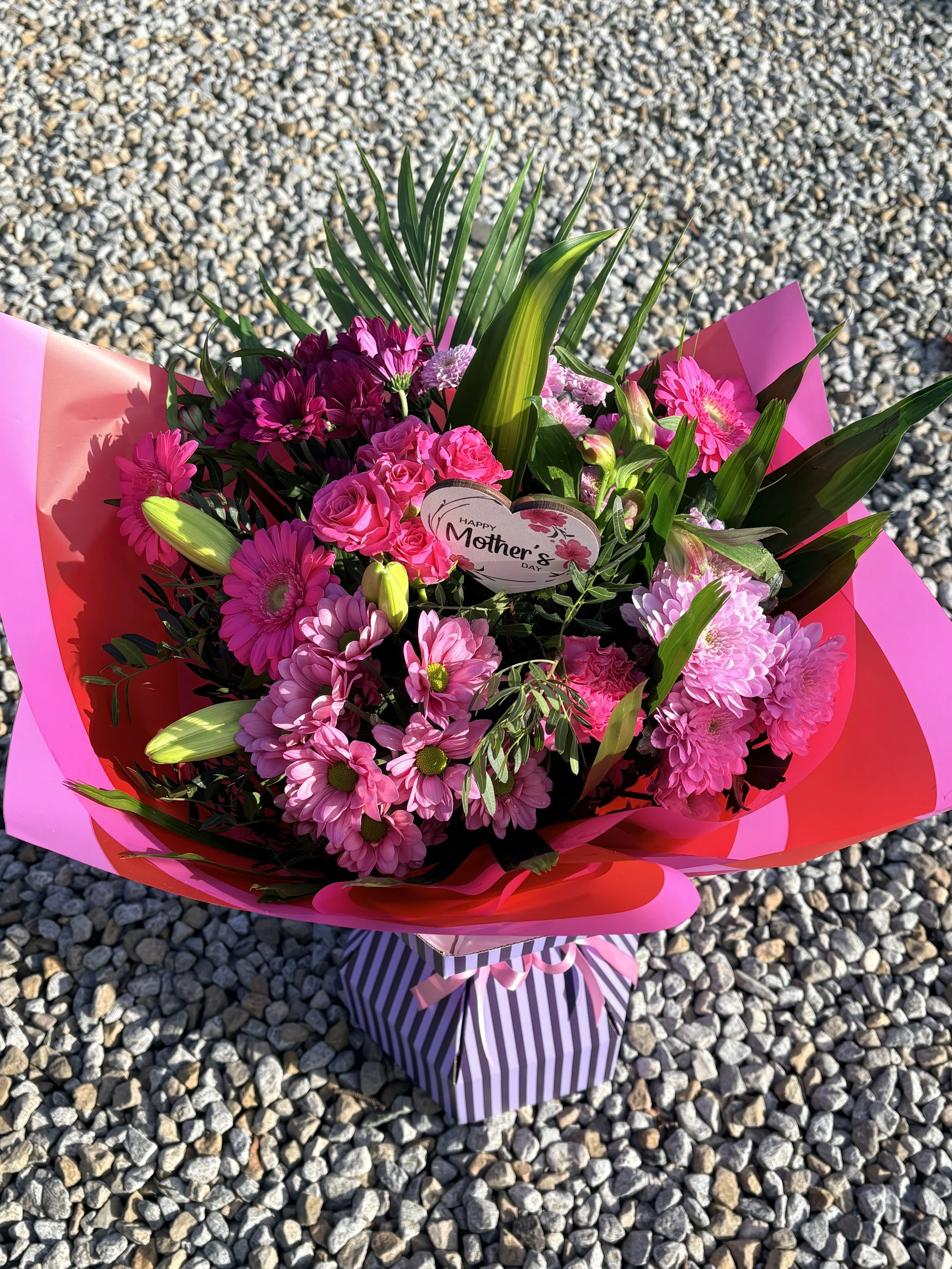 Bouquet of pink and purple flowers with green leaves, wrapped in pink and striped purple paper, with a tag that says "Happy Mother's Day." placed on a gravel surface.