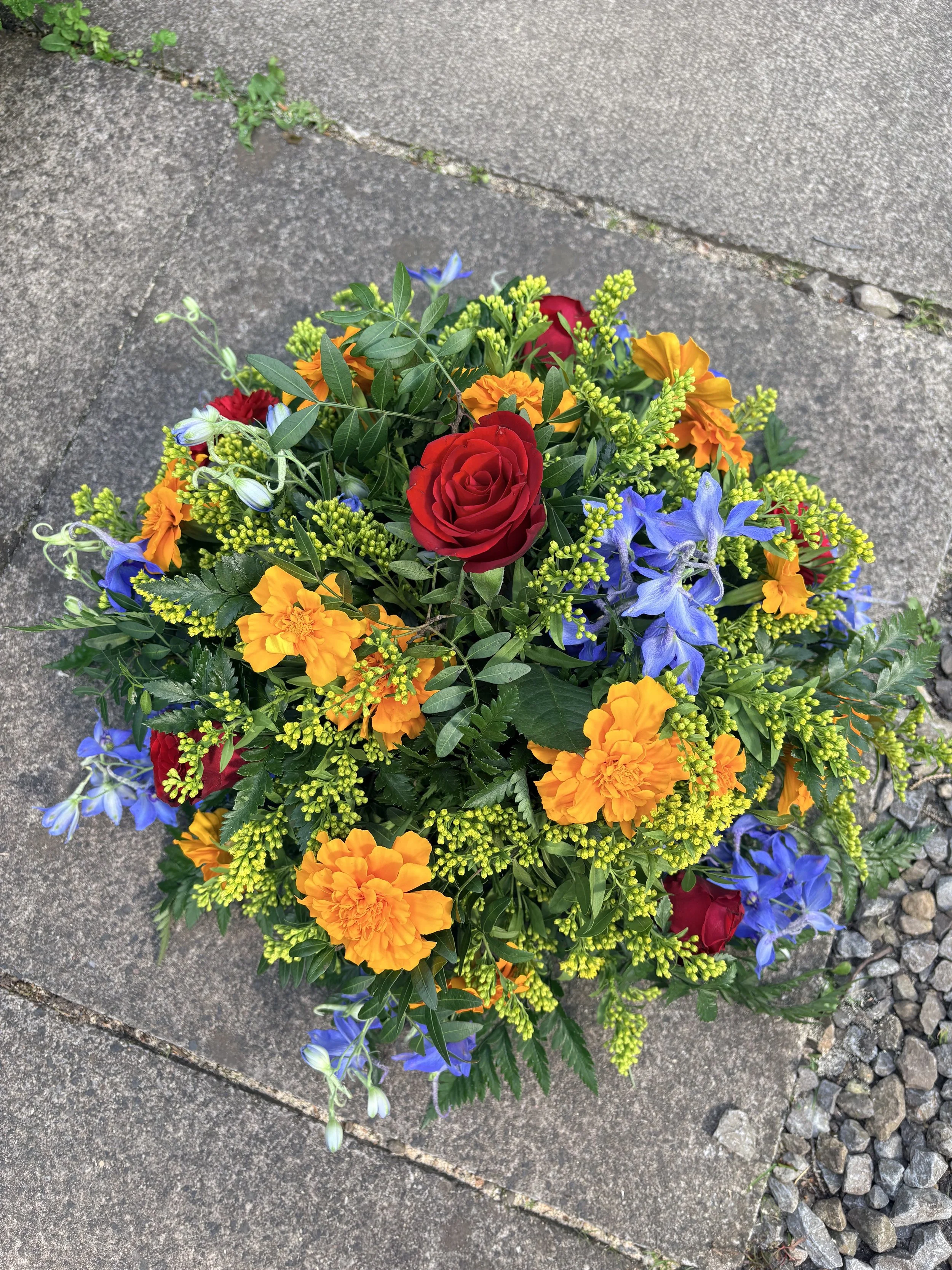 A colourful bouquet of flowers containing red roses, orange marigolds, blue delphiniums, and green foliage 