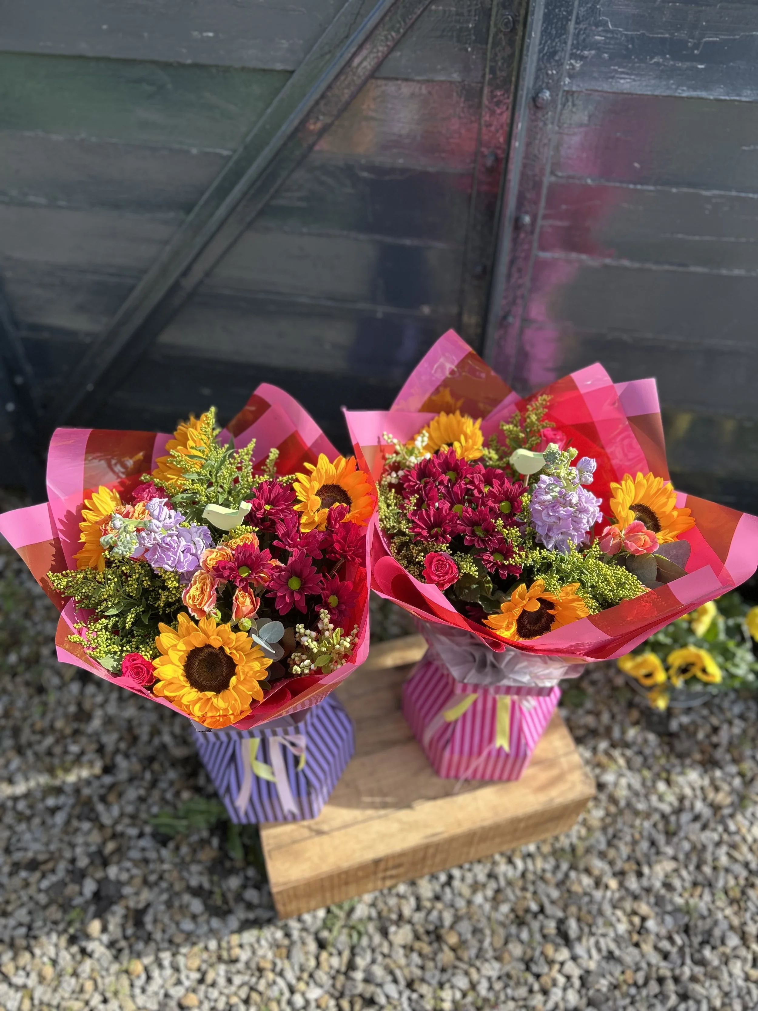 Two colorful bouquets of flowers with sunflowers, roses, and other blooms, wrapped in pink and purple striped paper, sitting on a wooden surface outside on a gravel ground.