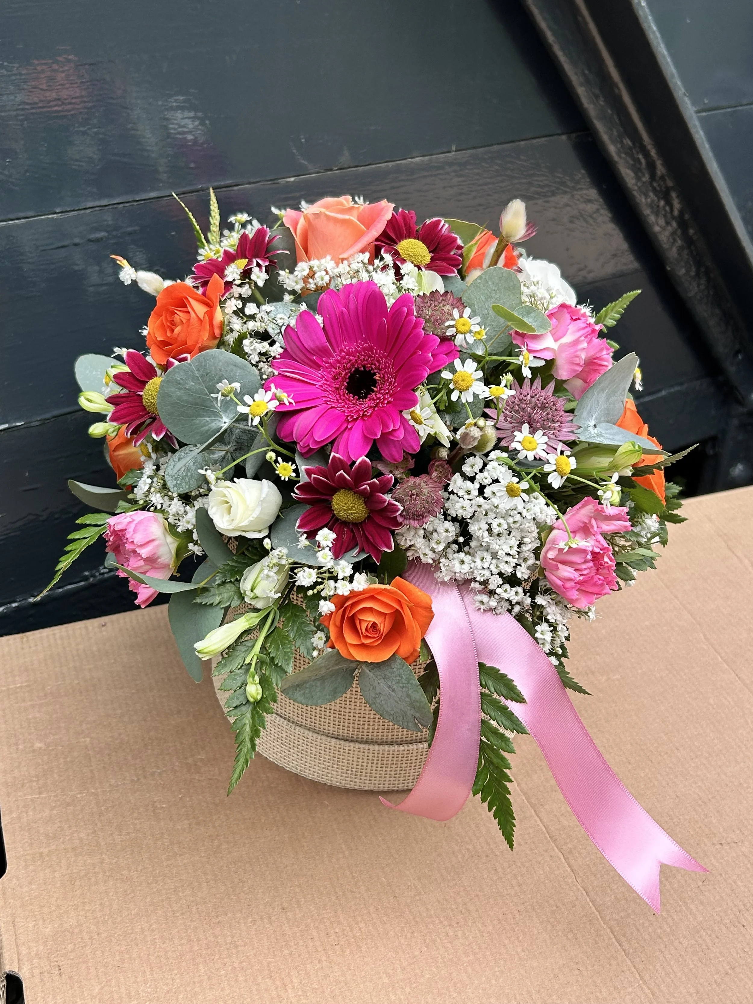 Colorful flower bouquet with pink gerbera daisy, orange roses, white and pink carnations, small white daisies, and greenery, arranged with a pink ribbon, on a brown paper-covered surface.
