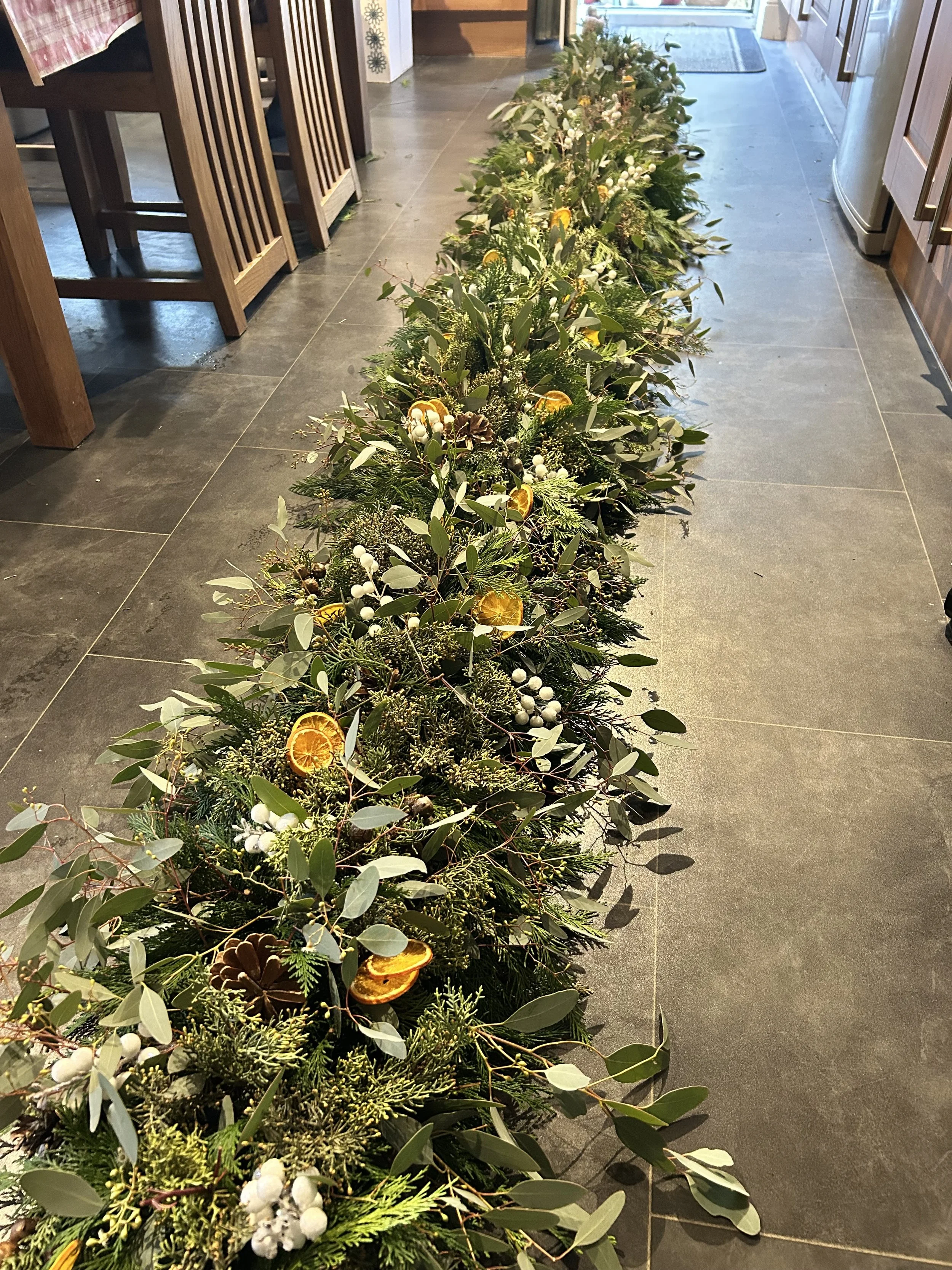 A long green holiday garland decorated with dried orange slices, pine cones, white berries, and various green foliage, placed on a kitchen floor near the entrance door.