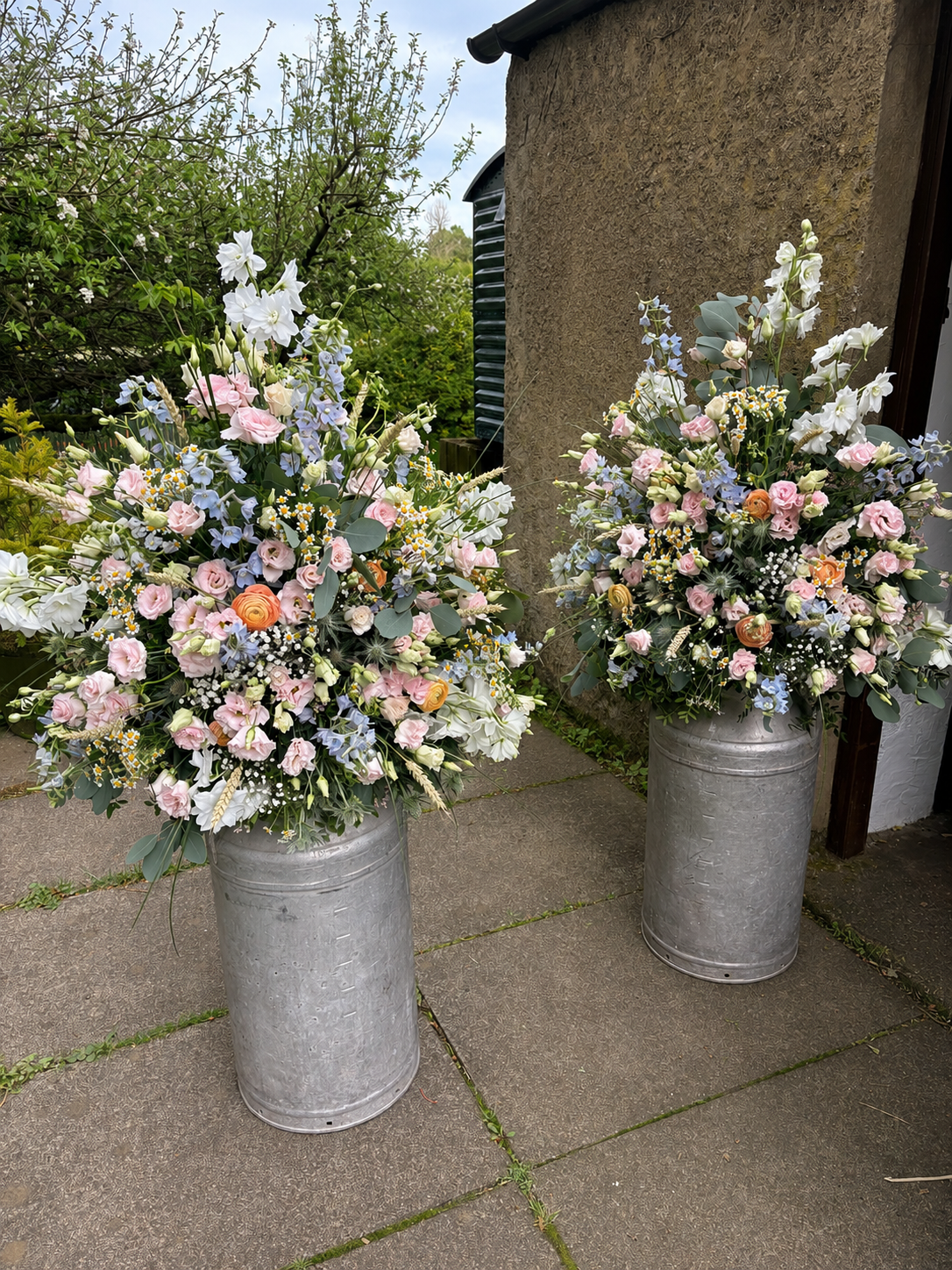 Two large metal planters filled with pastel-colored floral arrangements, including roses, lilies, and other flowers, outside on a tiled walkway with greenery and a building wall in the background.