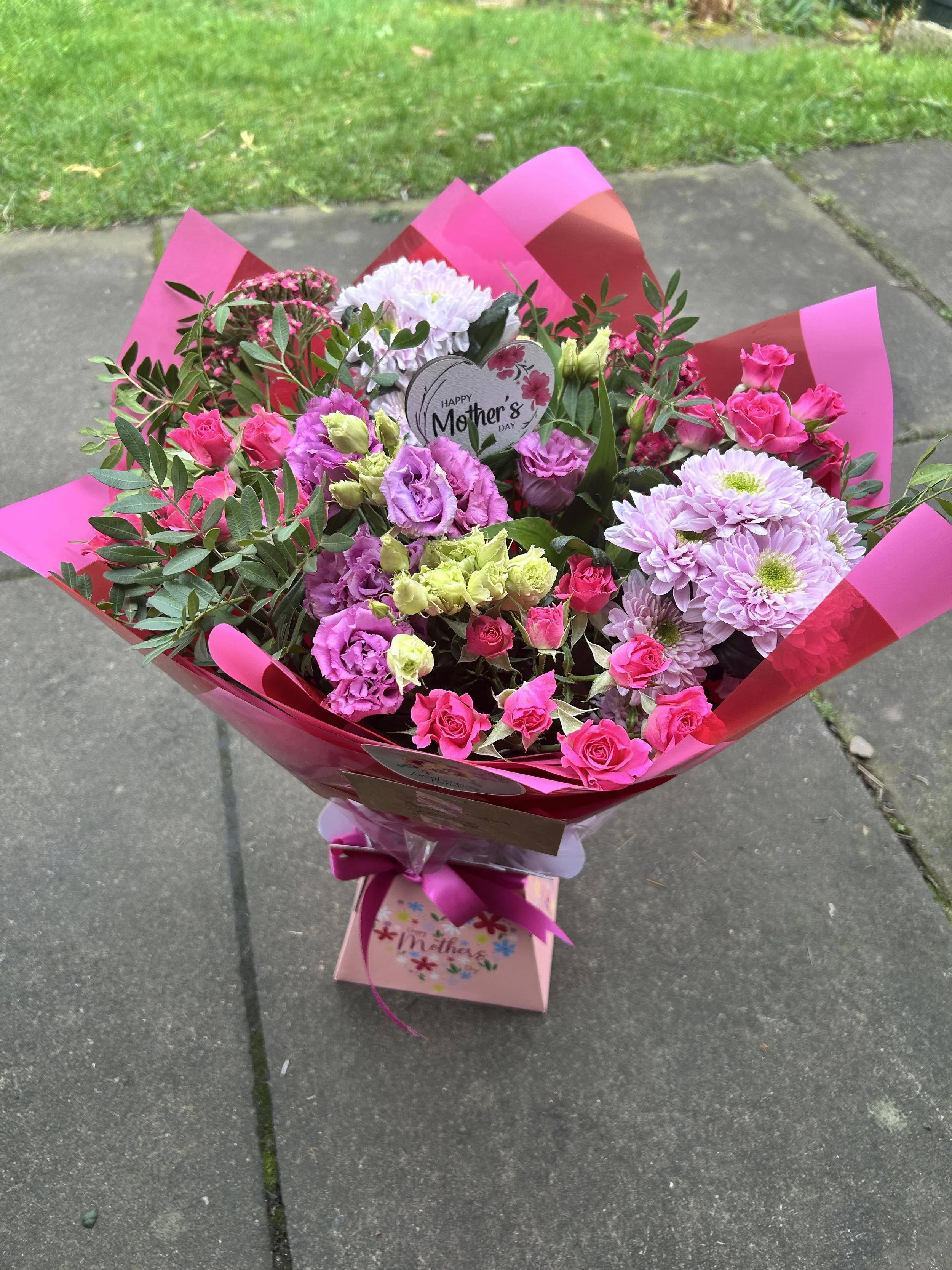 A bouquet of pink, purple, and white flowers with green leaves, wrapped in pink and red paper, and a sticker that says 'Happy Mother's Day', sitting on a concrete sidewalk.