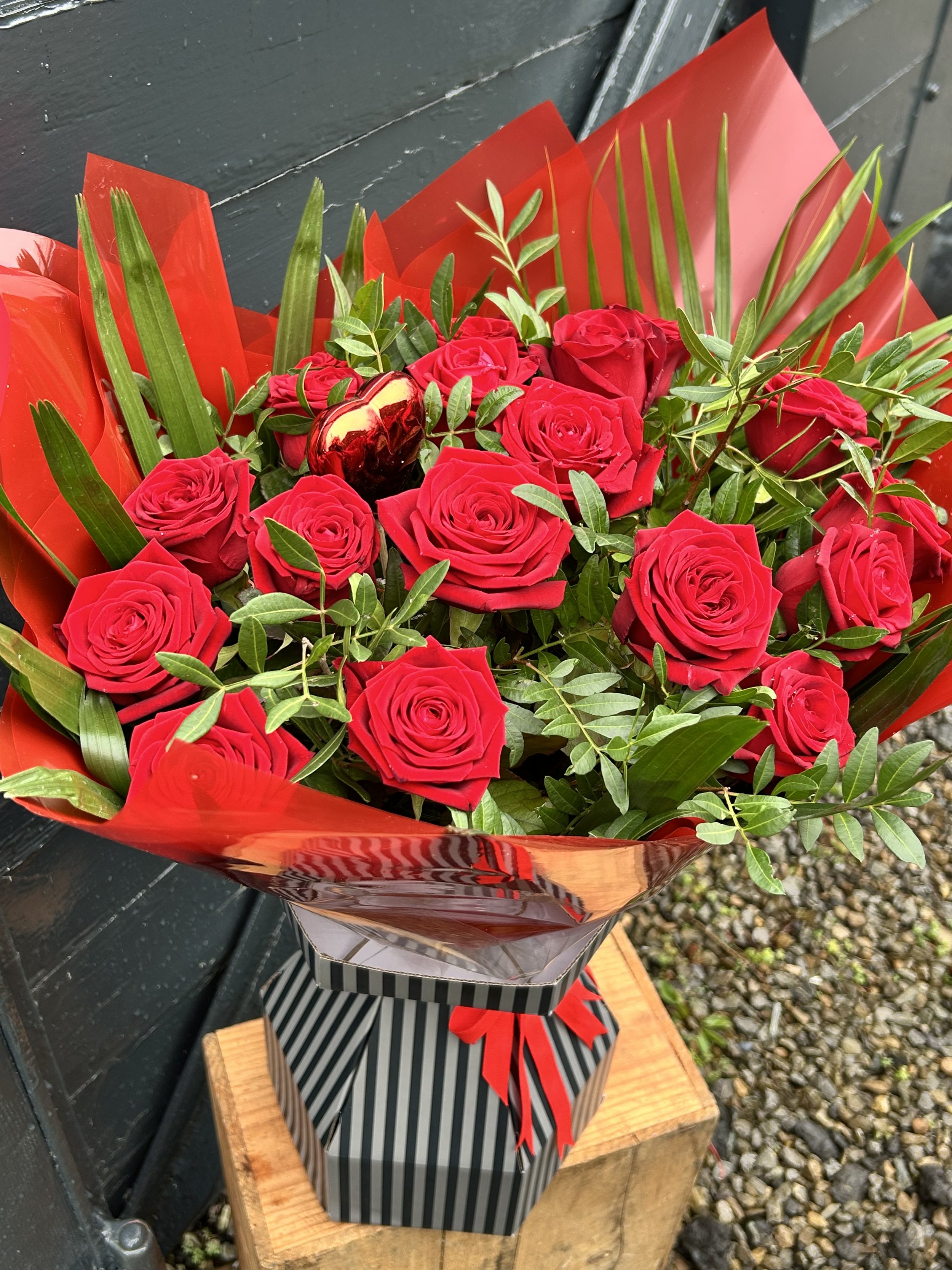 Bouquet of red roses wrapped in red and black striped and solid red wrapping paper, tied with a red ribbon, placed on a wooden stand outdoors.