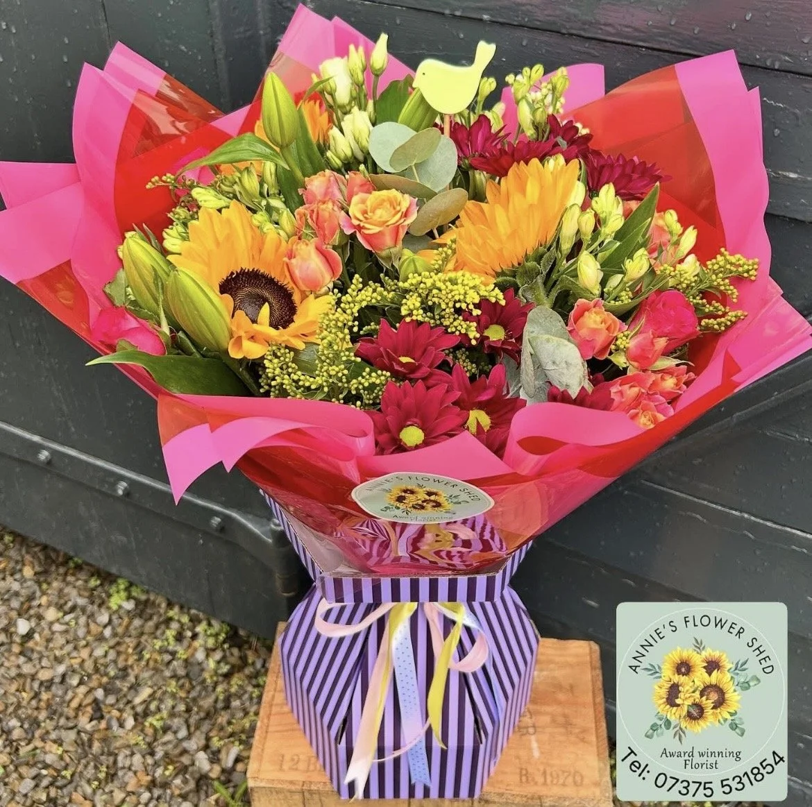 Bouquet of colorful mixed flowers, including sunflowers, roses, daisies, and greenery, wrapped in pink and red paper with a purple striped base, placed on a wooden surface outdoors.