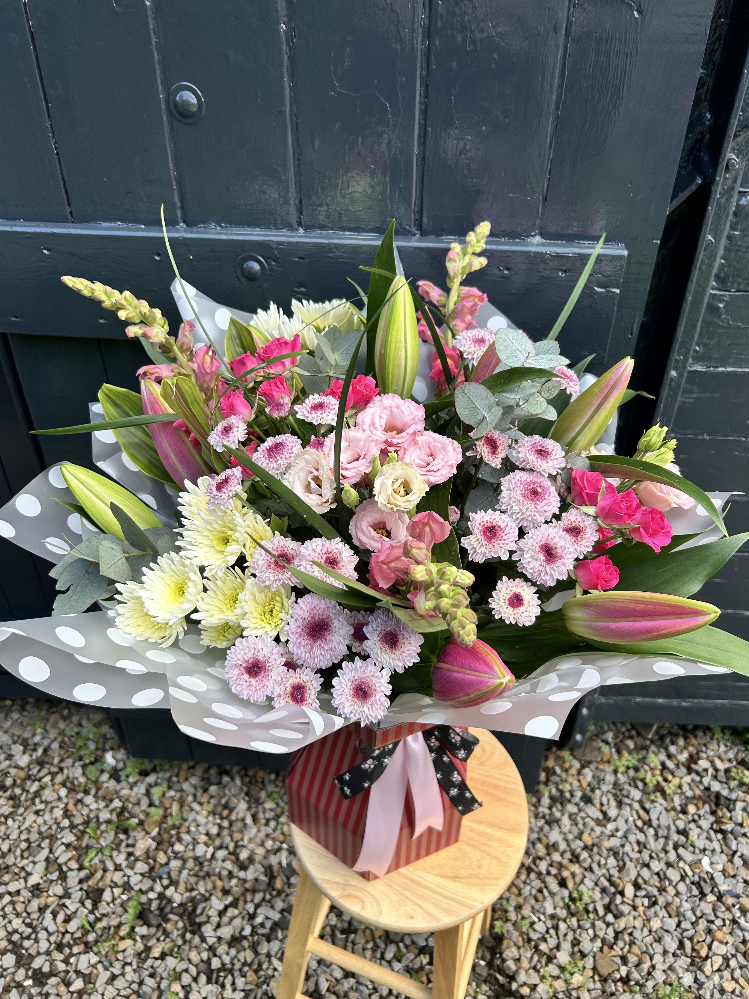 A bouquet of pink and white flowers, including lilies, carnations, and chrysanthemums, wrapped in decorative paper with pink and black ribbons, arranged on a small wooden stool outdoors with pebbled ground.