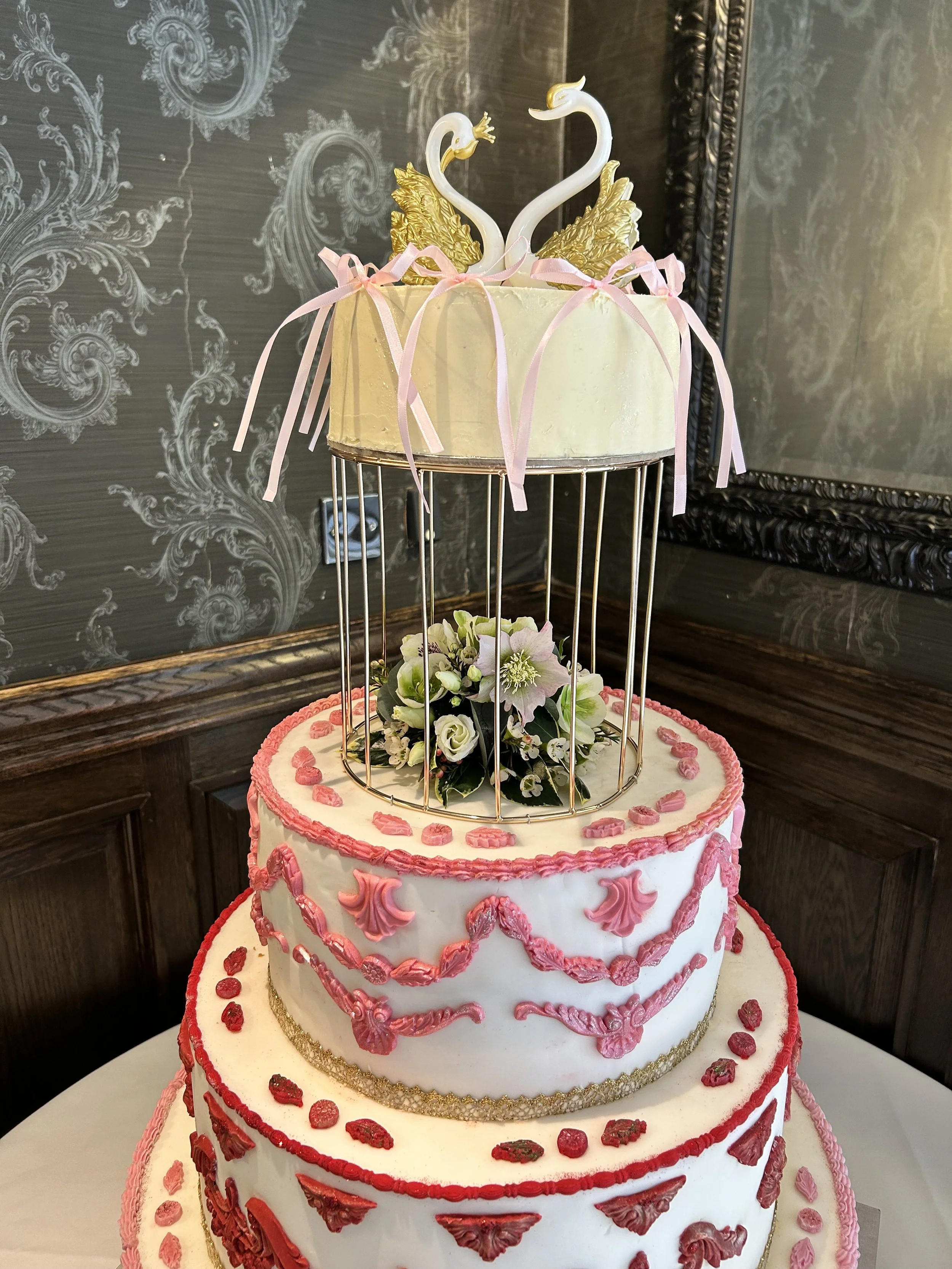 A multi-tiered wedding cake decorated with pink and red icing, topped with a gold swan and two white swans with gold accents, and adorned with a pink ribbon and fresh flowers.