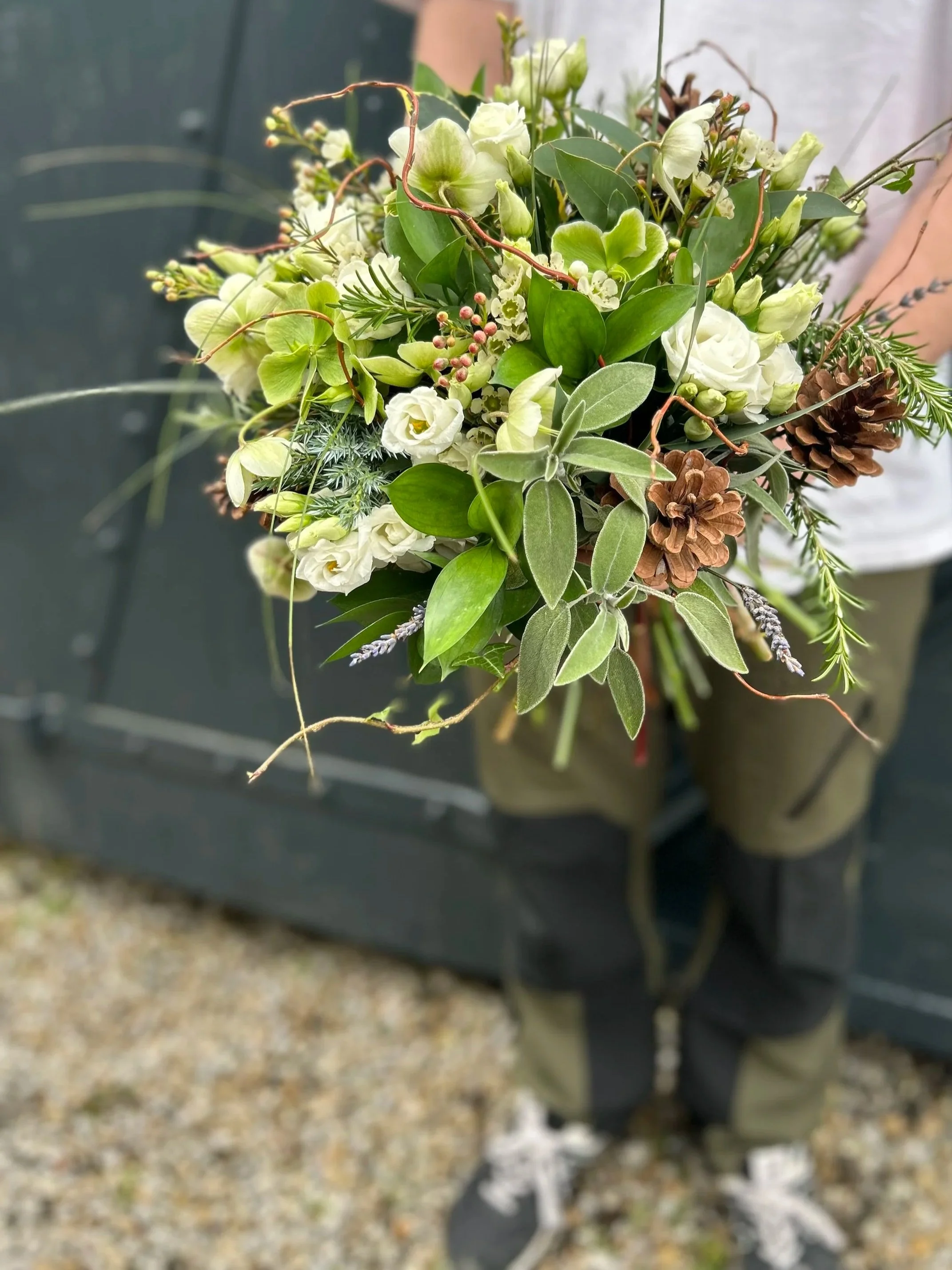 A bouquet of white and green flowers, surrounded by greenery and pinecones, held by a person wearing gray sneakers with white soles.